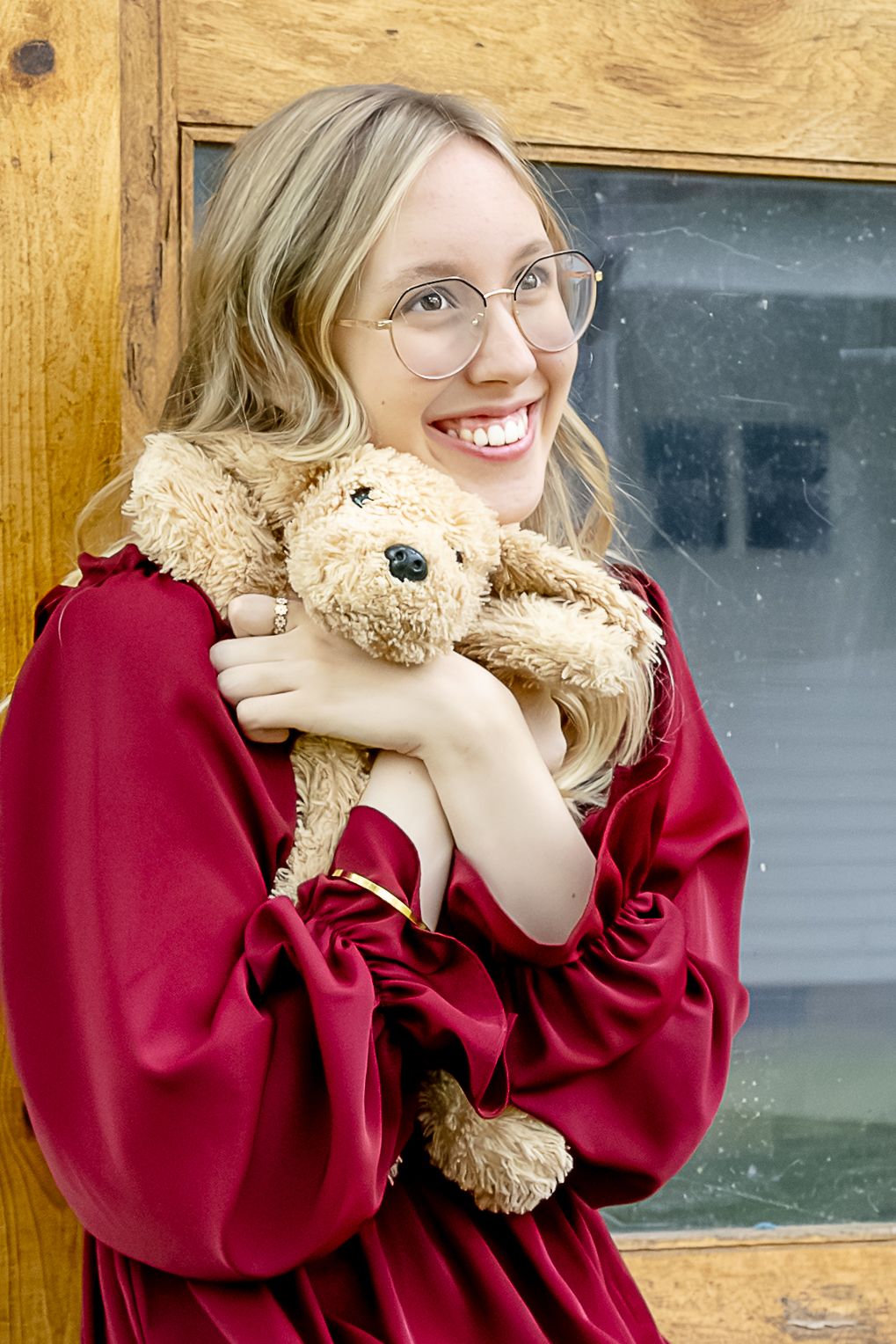Smiling blonde woman in red dress hugging a tan teddy bear, wearing glasses, leaning against a wooden door.