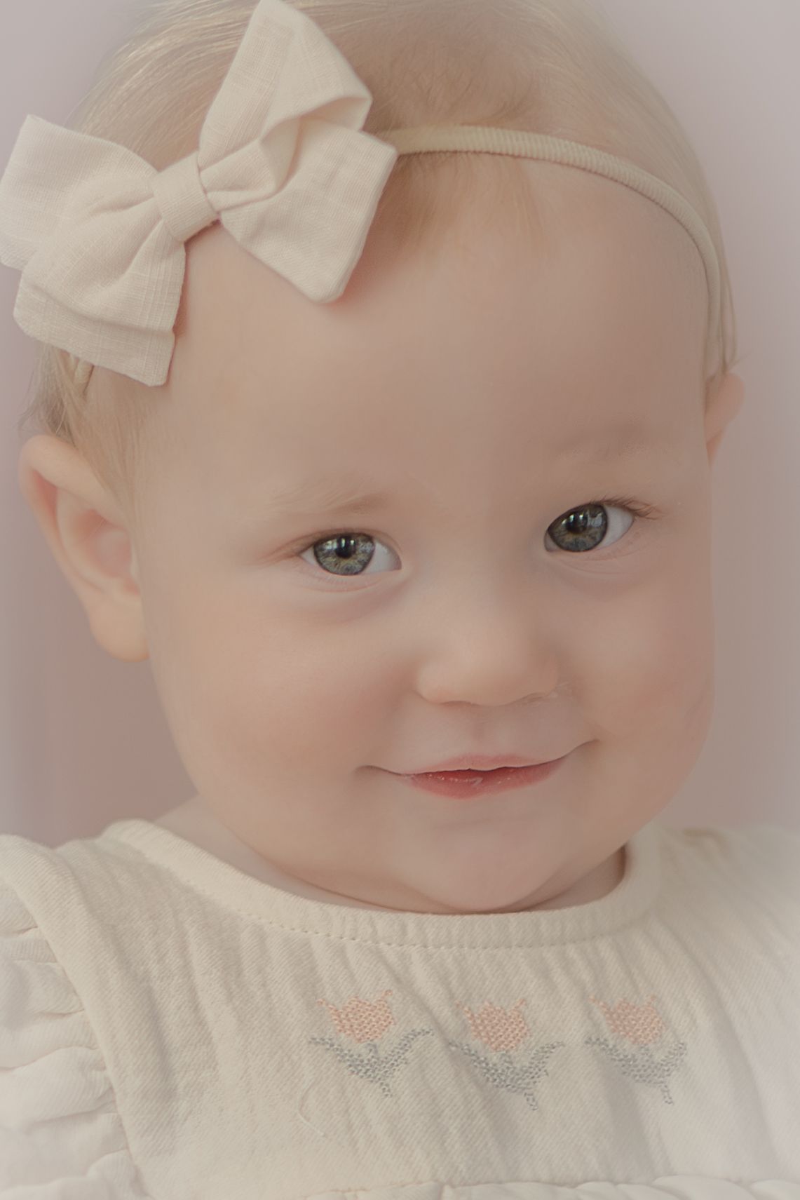 Adorable baby girl with blue eyes wearing a white bow headband and embroidered dress, smiling softly.