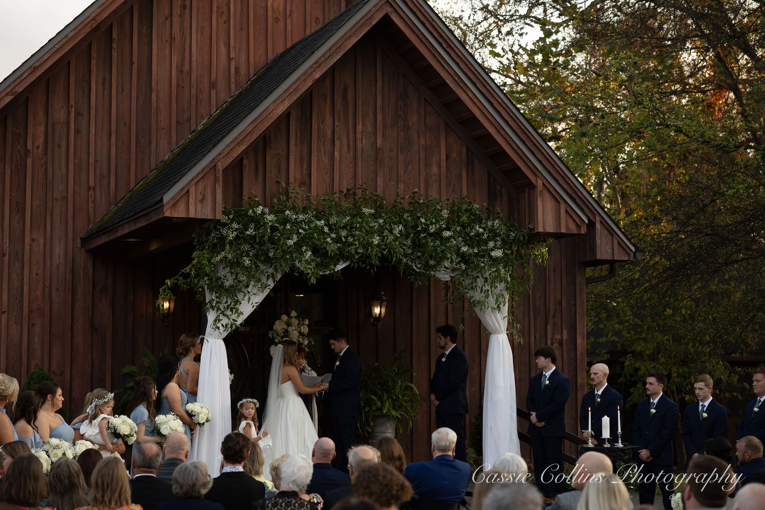 Wedding ceremony taking place in a rustic wooden barn venue with string lights and white fabric draping the ceiling.