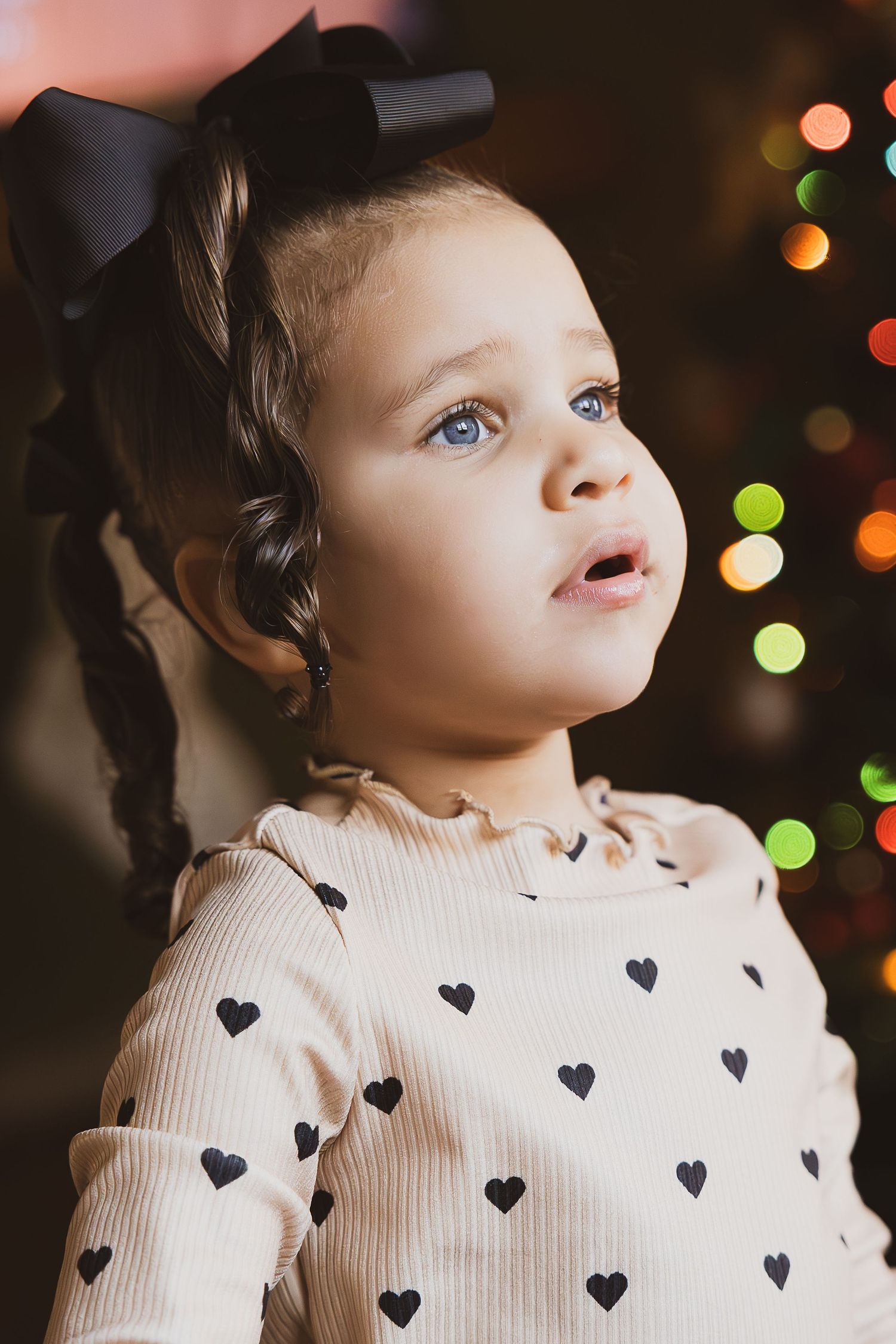 A young child in a cream sweater with heart patterns gazes upward while standing against a blurred background of holiday lights.