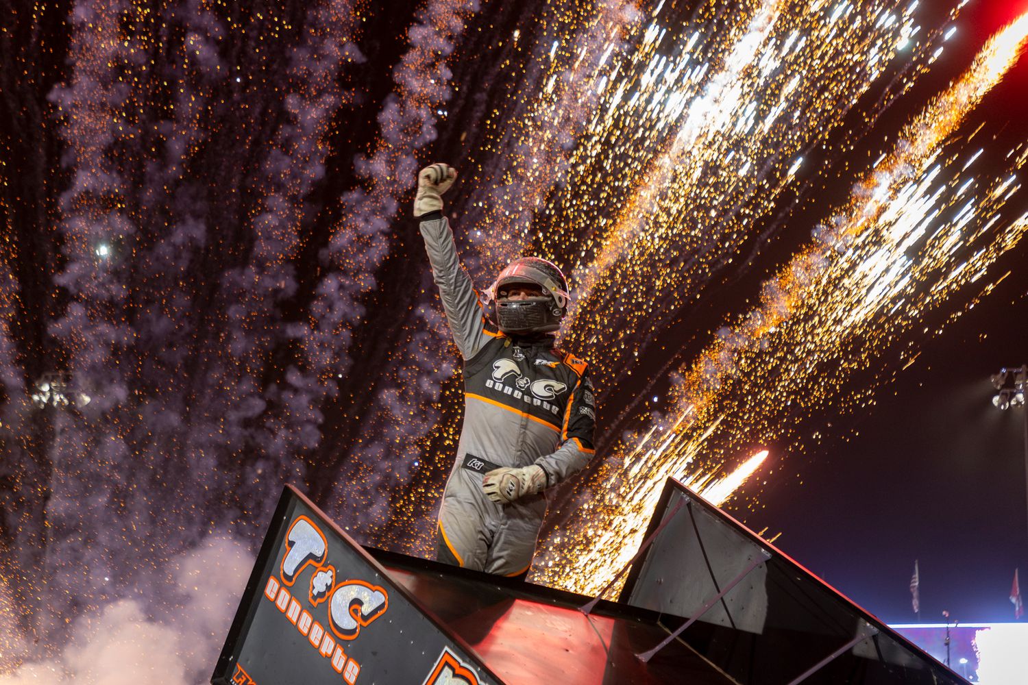 Sprint car driver raises a fist in victory as fireworks explode from the top wing at Skagit Speedway.