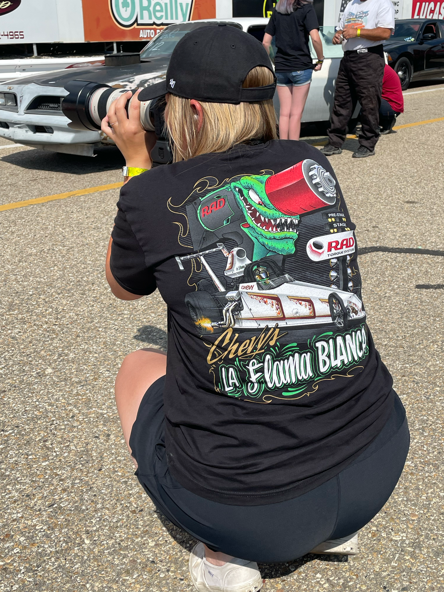 Becky squats low with a long lens, repping a Chevy Reeves Racing tee while capturing the action at the track.