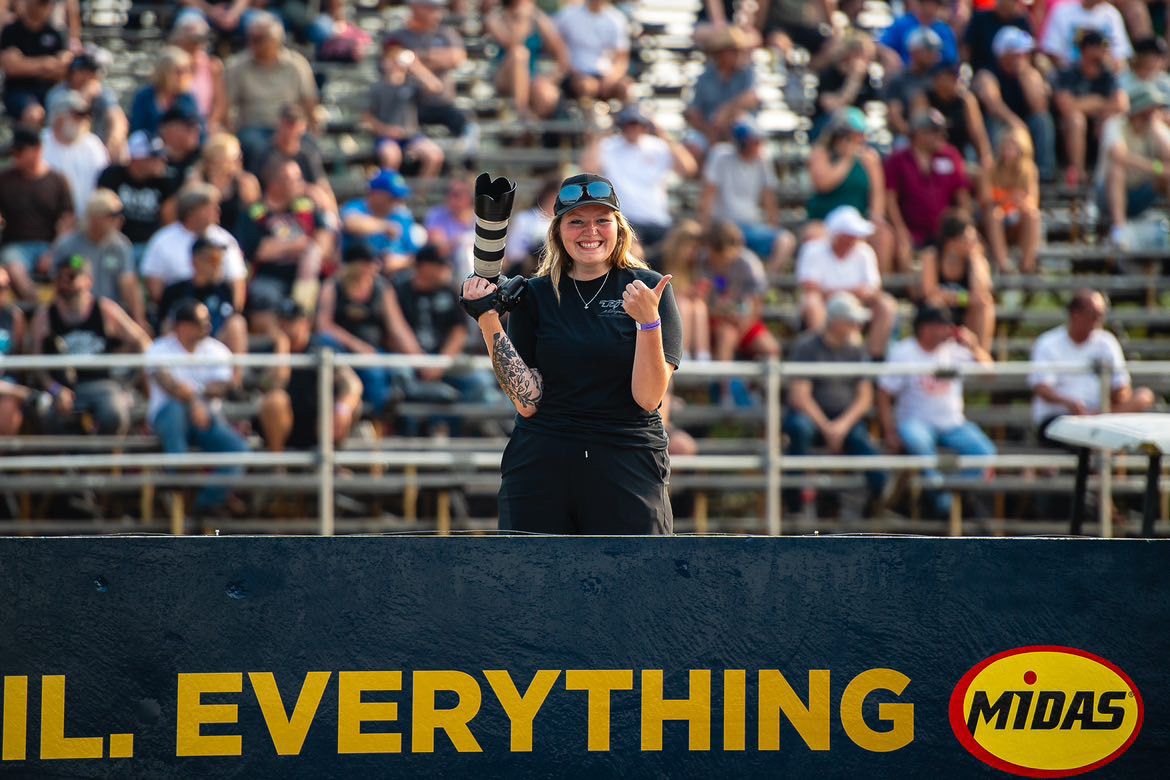 Becky smiling with camera in hand, standing in front of a packed grandstand during a motorsports event.