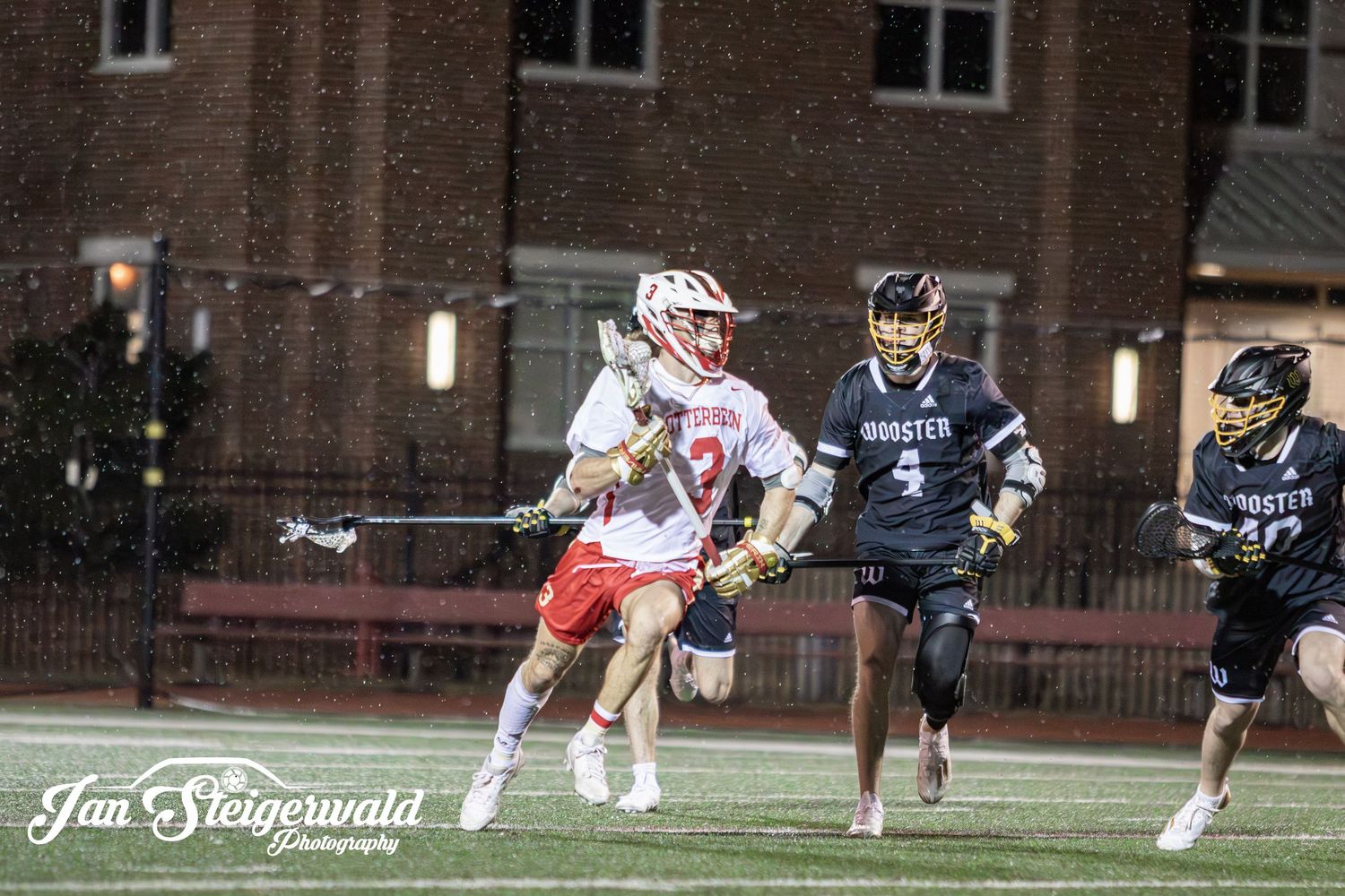 Lacrosse players compete in a nighttime match on an illuminated field with brick buildings in the background.