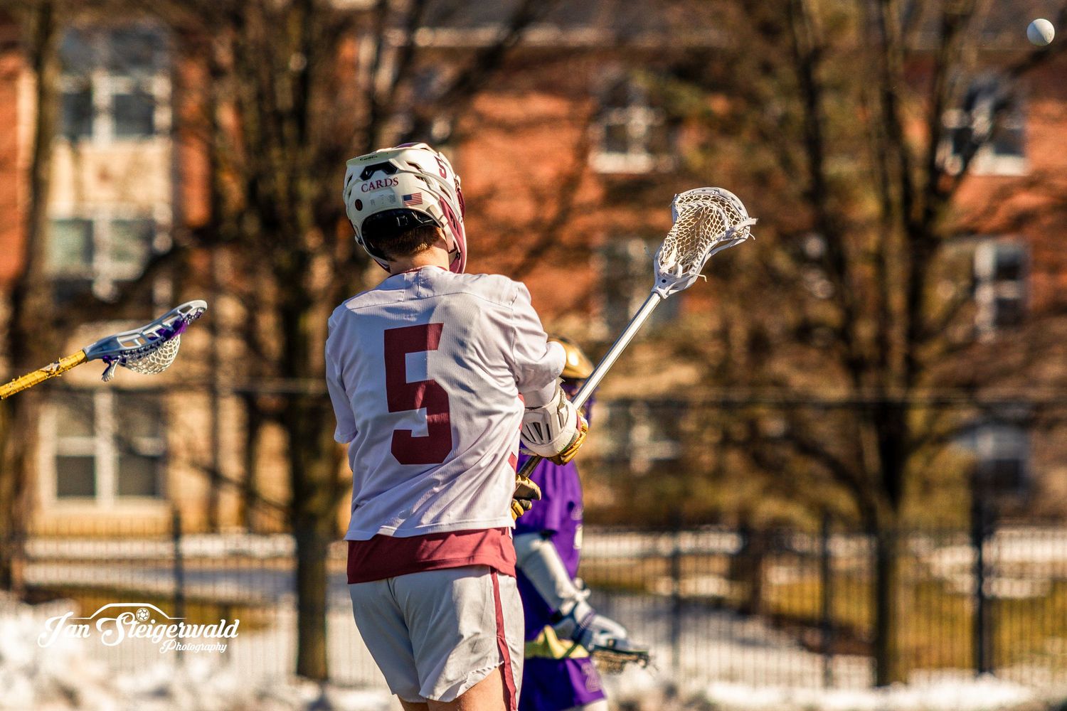 College lacrosse player in white jersey holding stick during game against purple team on snowy field.