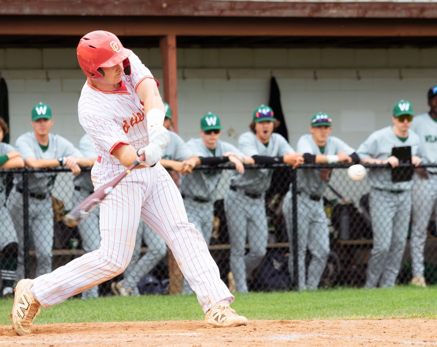 Baseball player in white pinstriped uniform swings bat while opposing team watches from dugout.