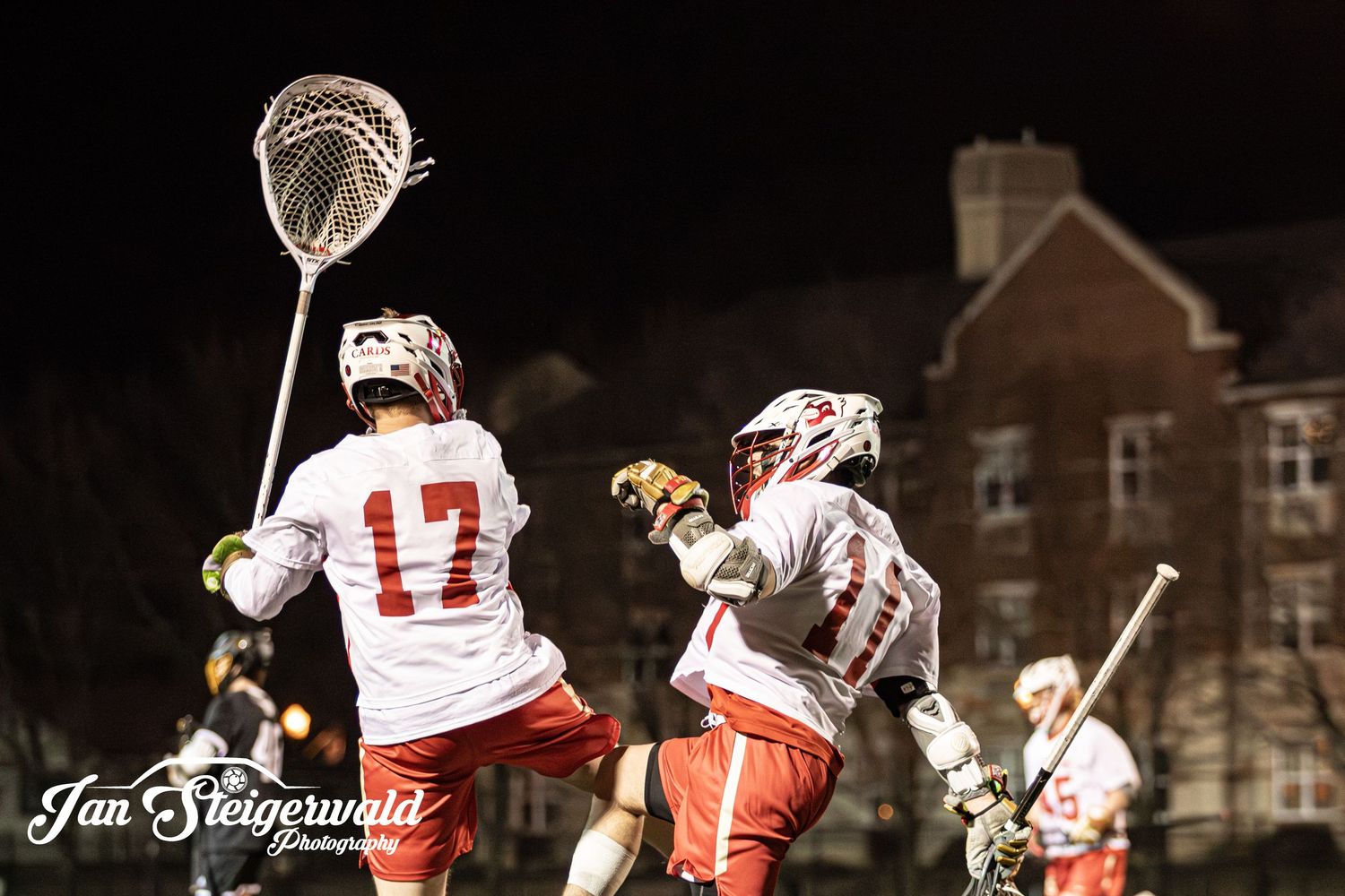 Lacrosse players in white and red uniforms compete at night with illuminated buildings visible in the background.