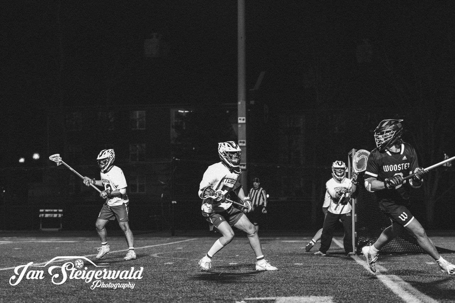 Black and white photo of lacrosse players in action during a nighttime game under stadium lights.