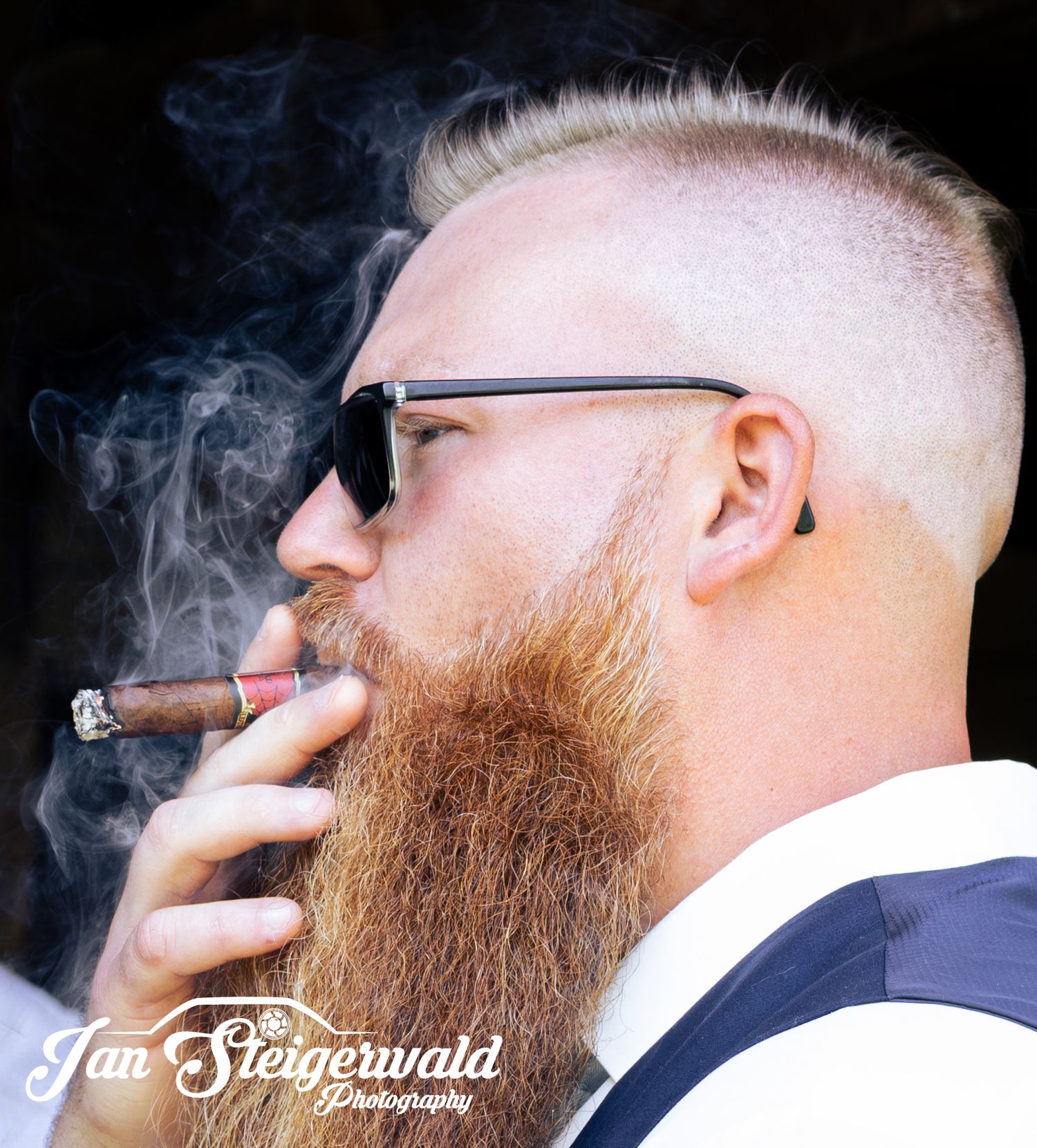 Side profile of someone with glasses and a long beard smoking a cigar with smoke rising against dark background.
