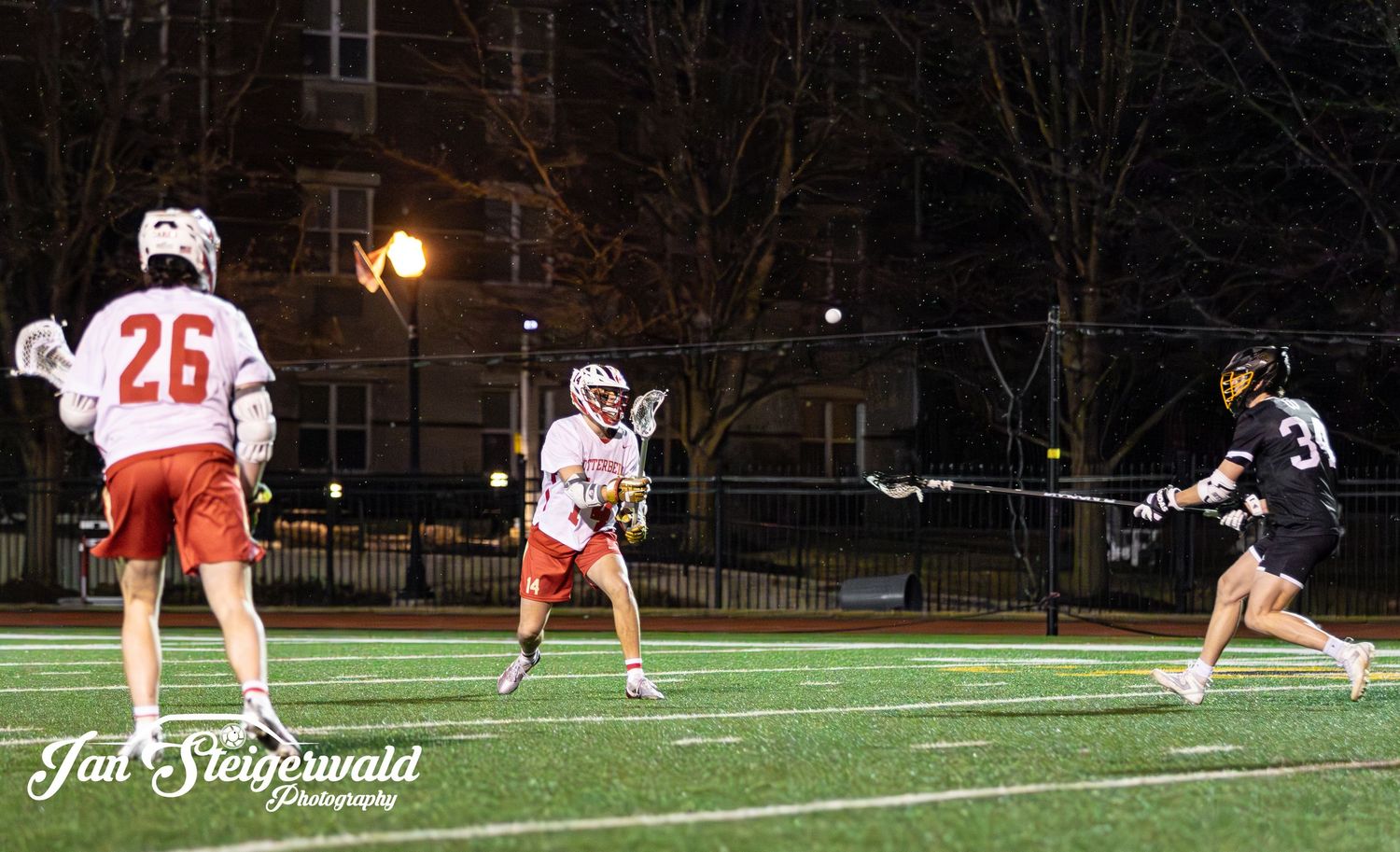 Three lacrosse players compete on an illuminated outdoor field at night during a match.