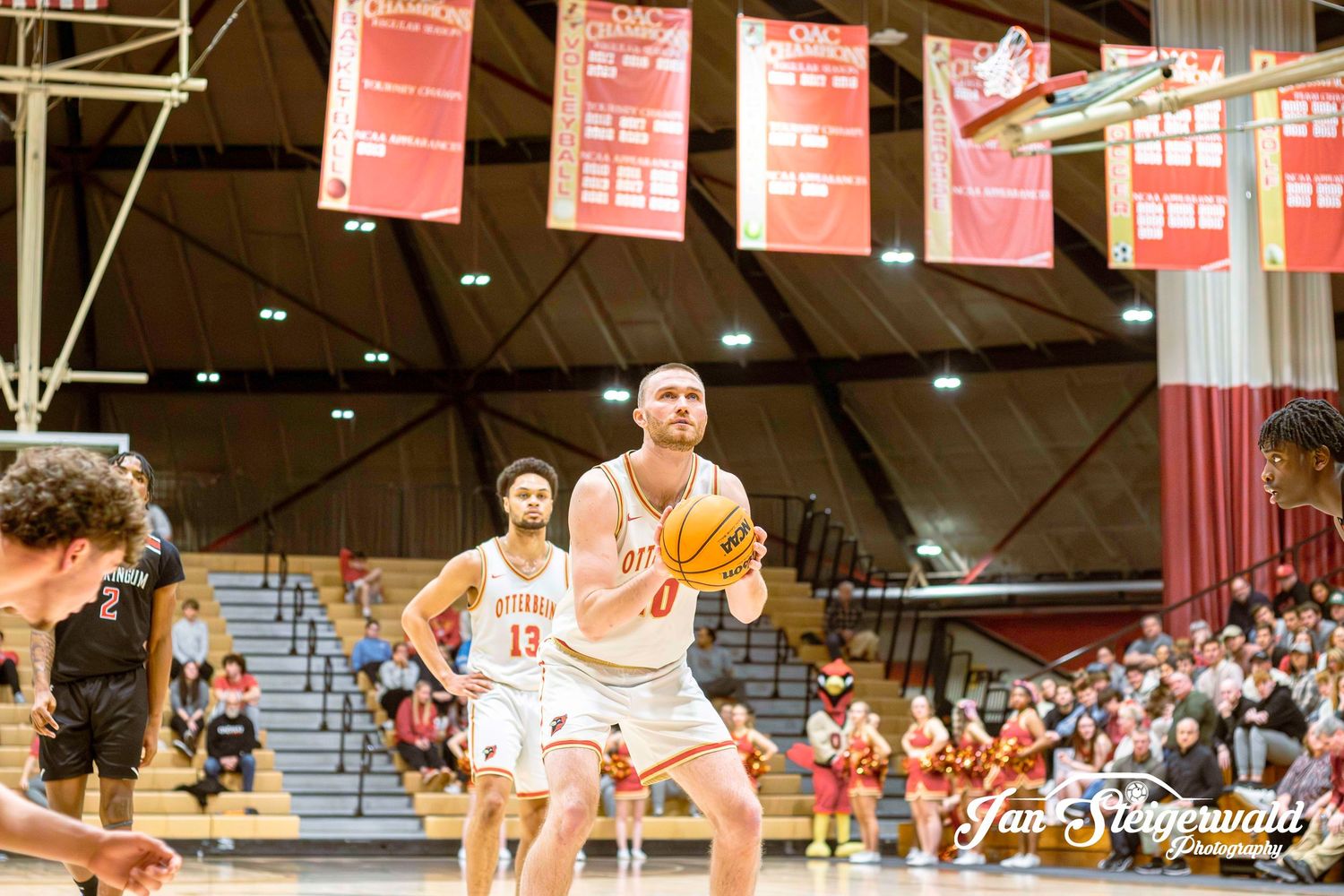 Players compete on a basketball court during an intense sporting match with red banners visible in the background.