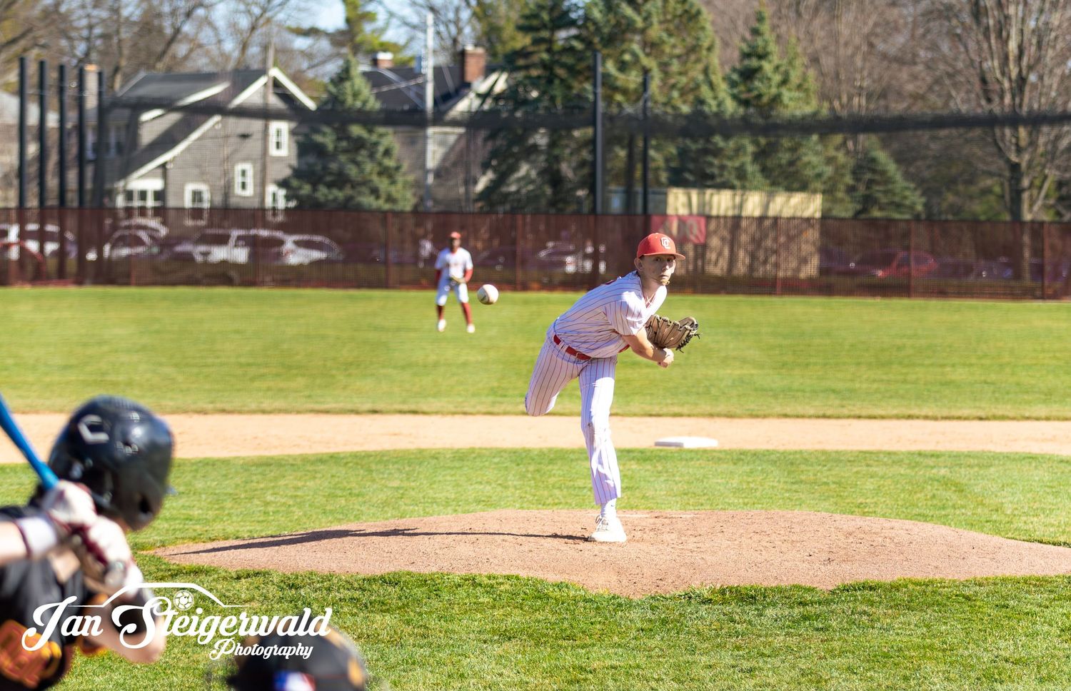 Baseball pitcher in purple jersey winds up to throw from the mound during a sunny afternoon game on grass field.