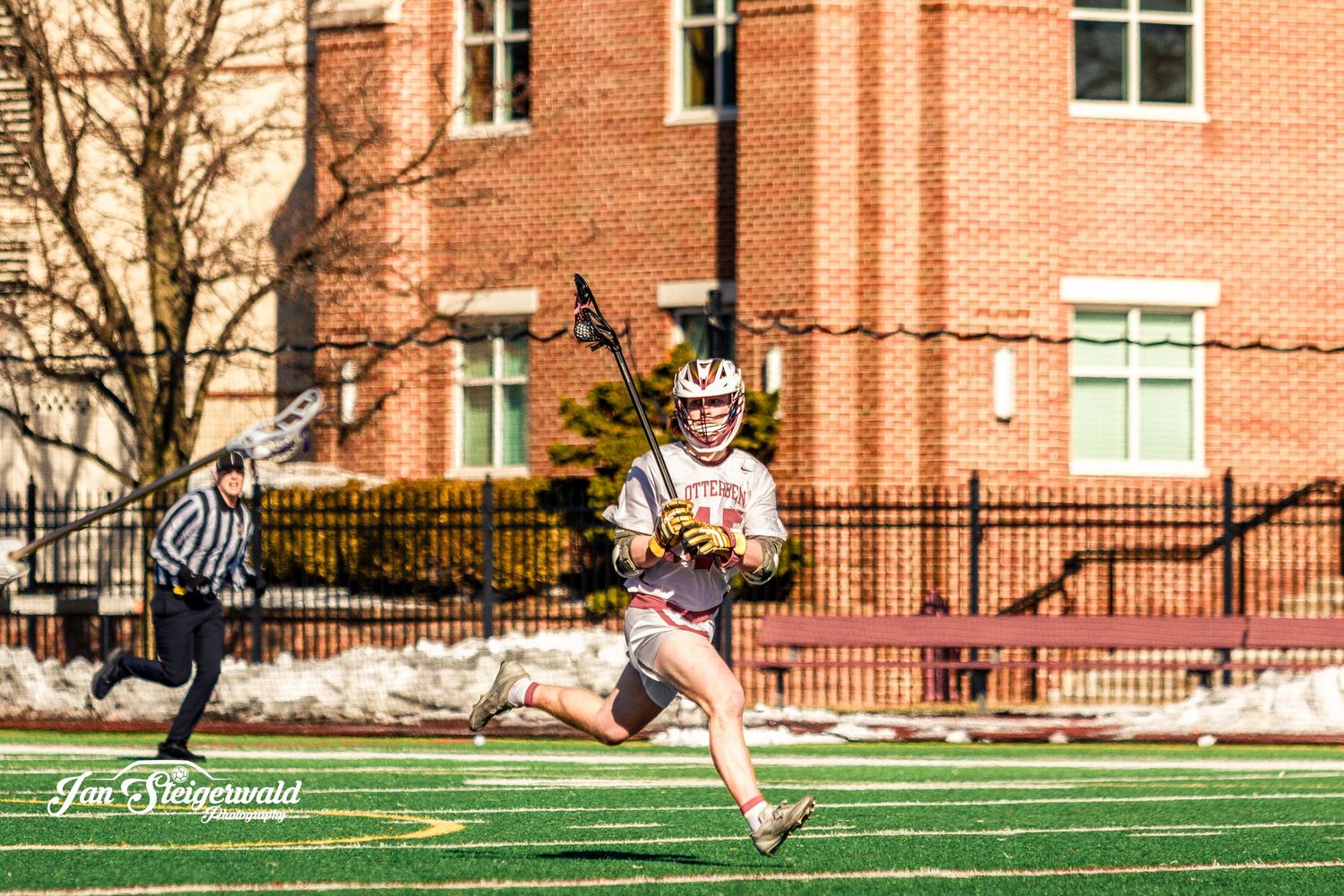 Lacrosse player in white uniform runs across field with stick during game with brick building in background.