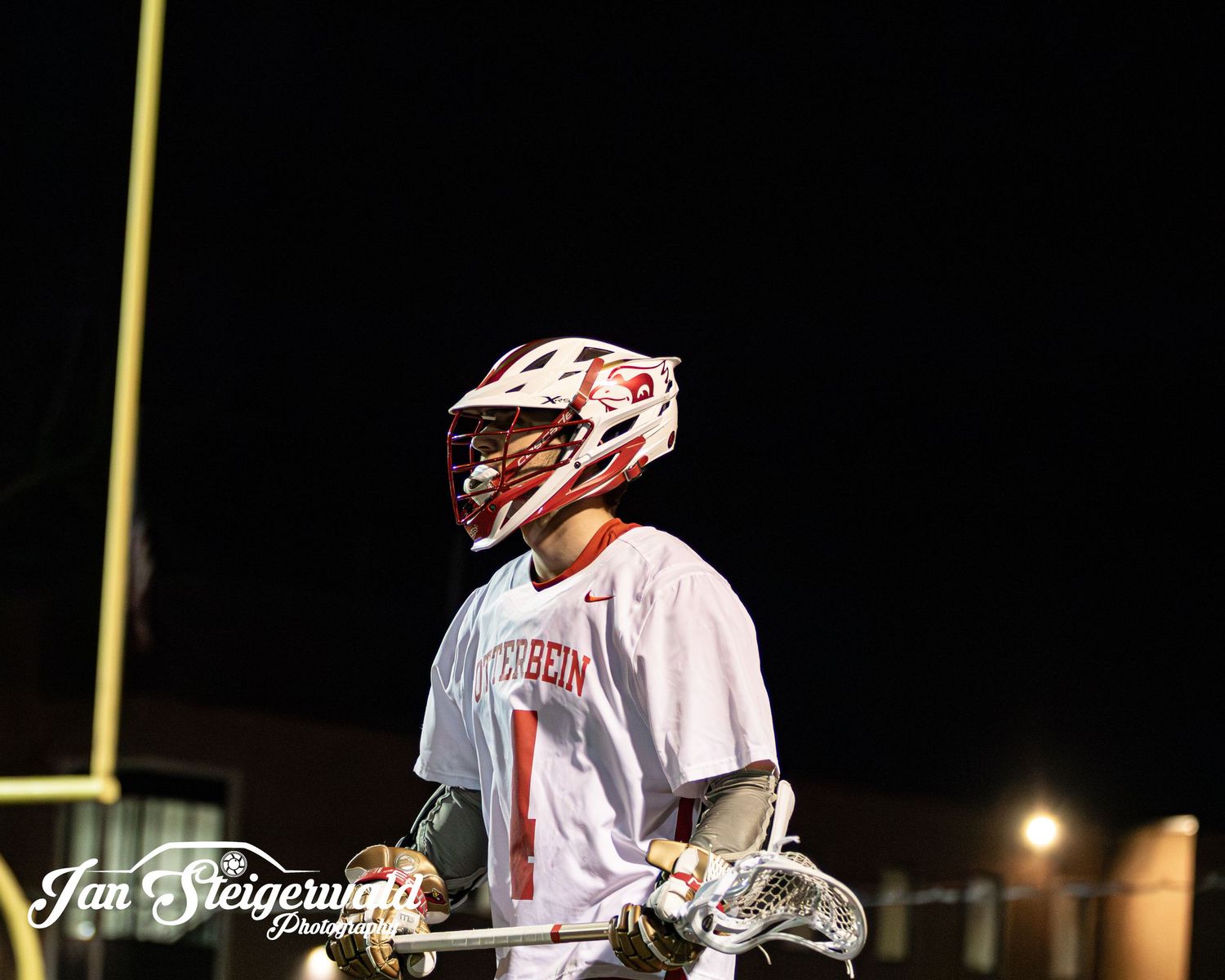 Lacrosse player in white uniform and red helmet stands on illuminated field at night during game.
