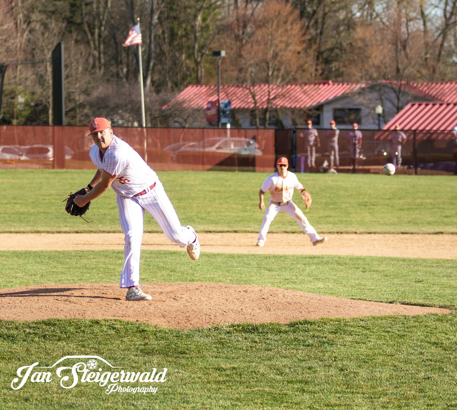 Baseball pitcher following through on delivery from dirt mound during sunny afternoon game on grass field with red dugout.