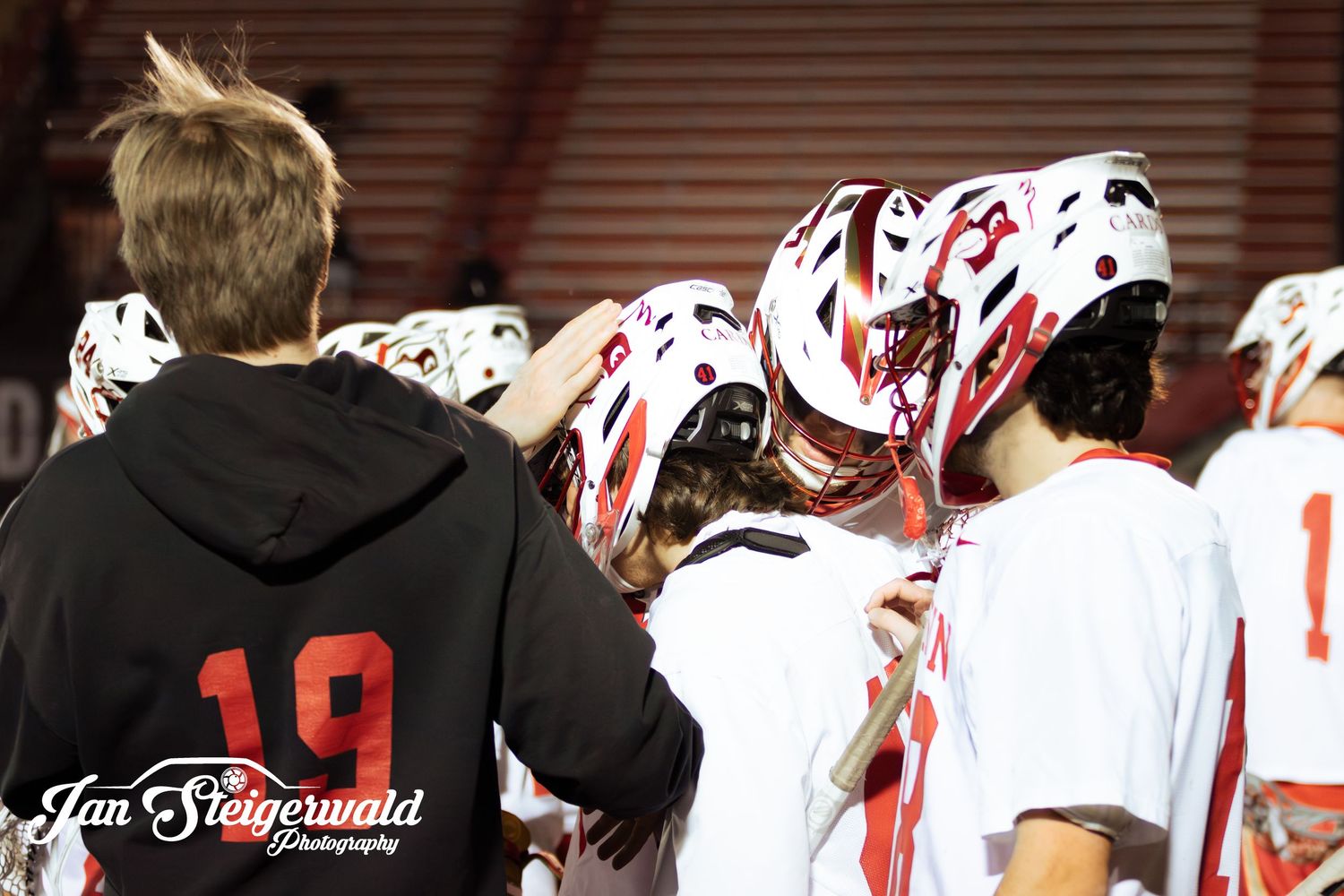 A group of lacrosse players wearing white uniforms with red accents huddle together during a game.
