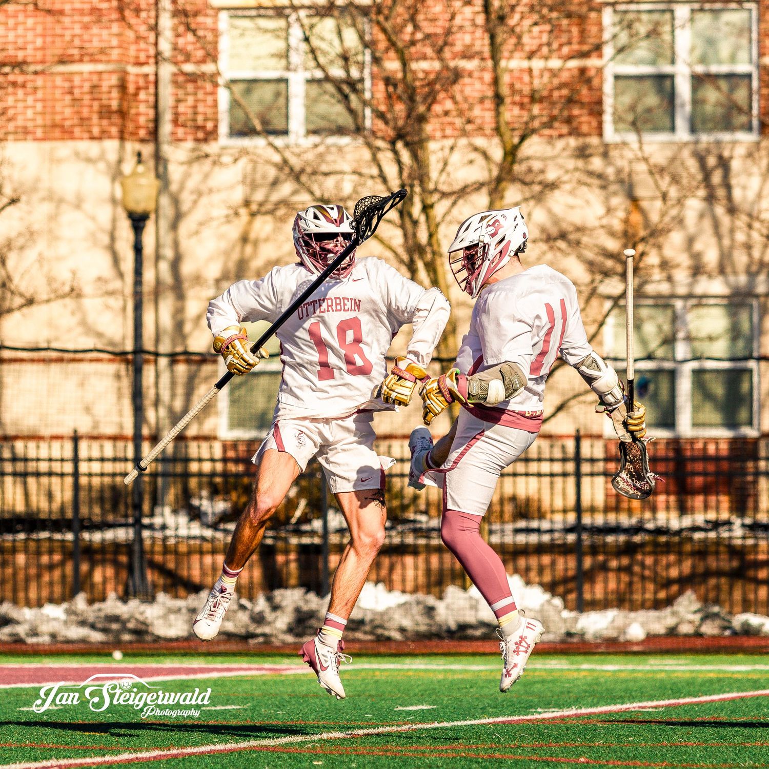 Two lacrosse players in white uniforms jump while playing on a field with brick buildings and bare trees in the background.