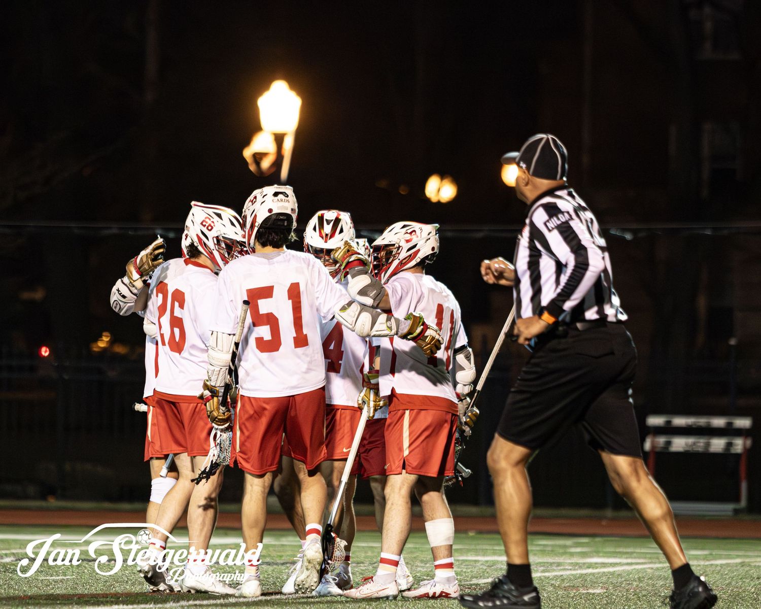 A lacrosse team huddles on a field at night under illuminated lamp posts as a referee approaches.