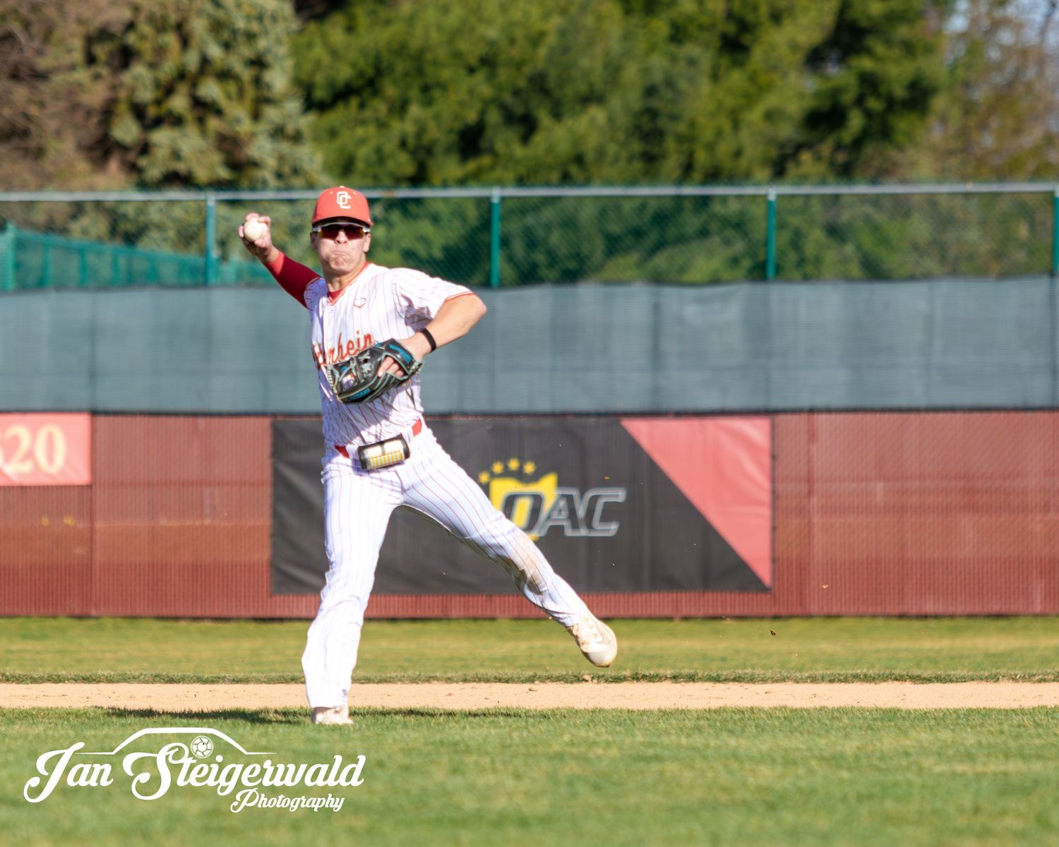 A panoramic view of a baseball field with red outfield wall and green trees in the background on a sunny day.