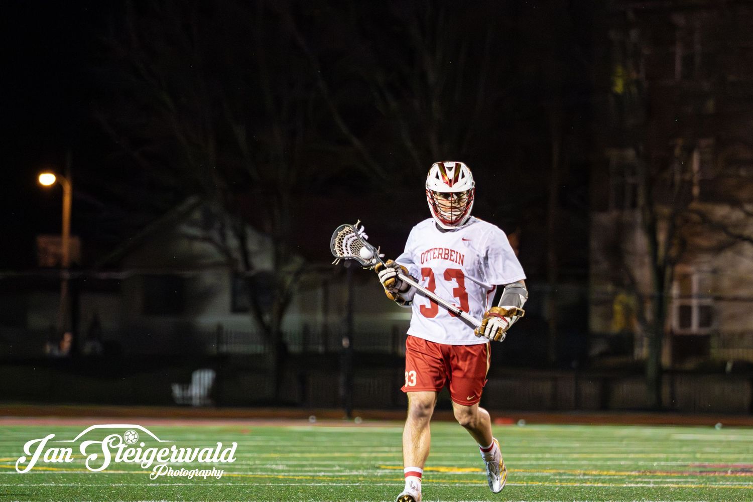 Lacrosse player in white and red uniform holds stick while playing under stadium lights at night.