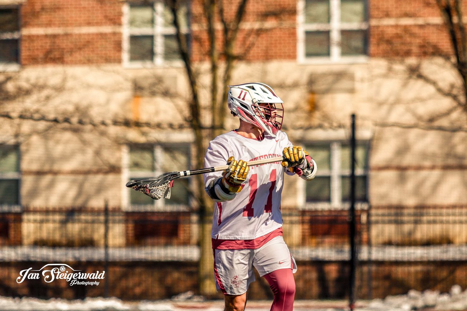 Lacrosse player in white jersey practices stick handling during outdoor training session at brick building complex.