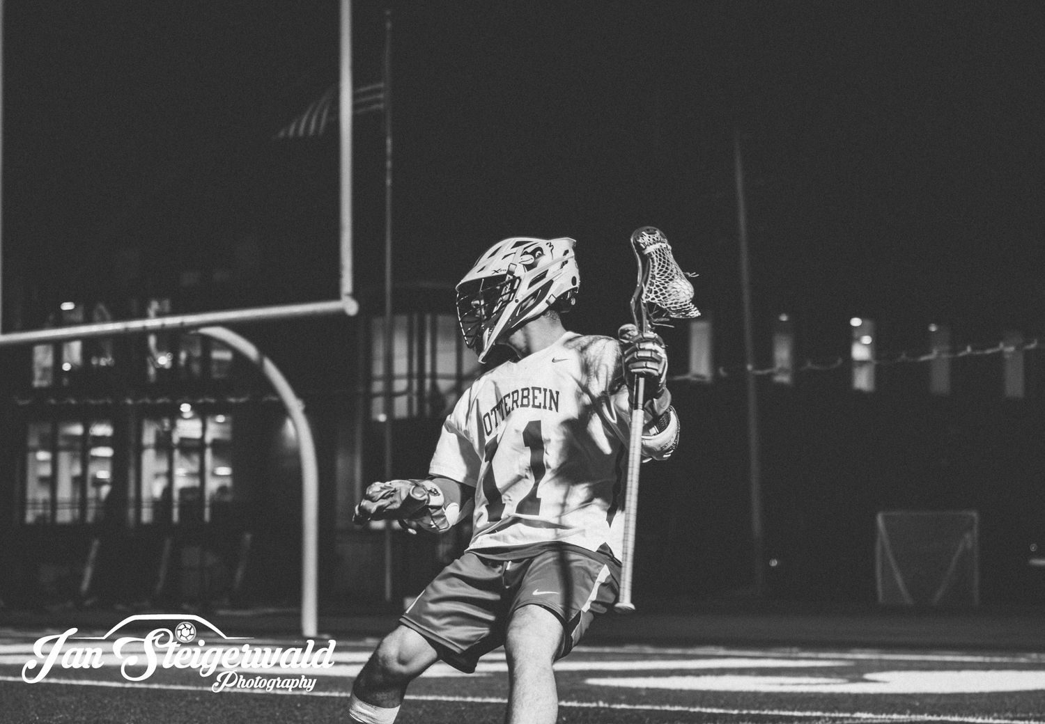 A lacrosse player in black and white photo follows through on a shot during a nighttime game on an outdoor field.