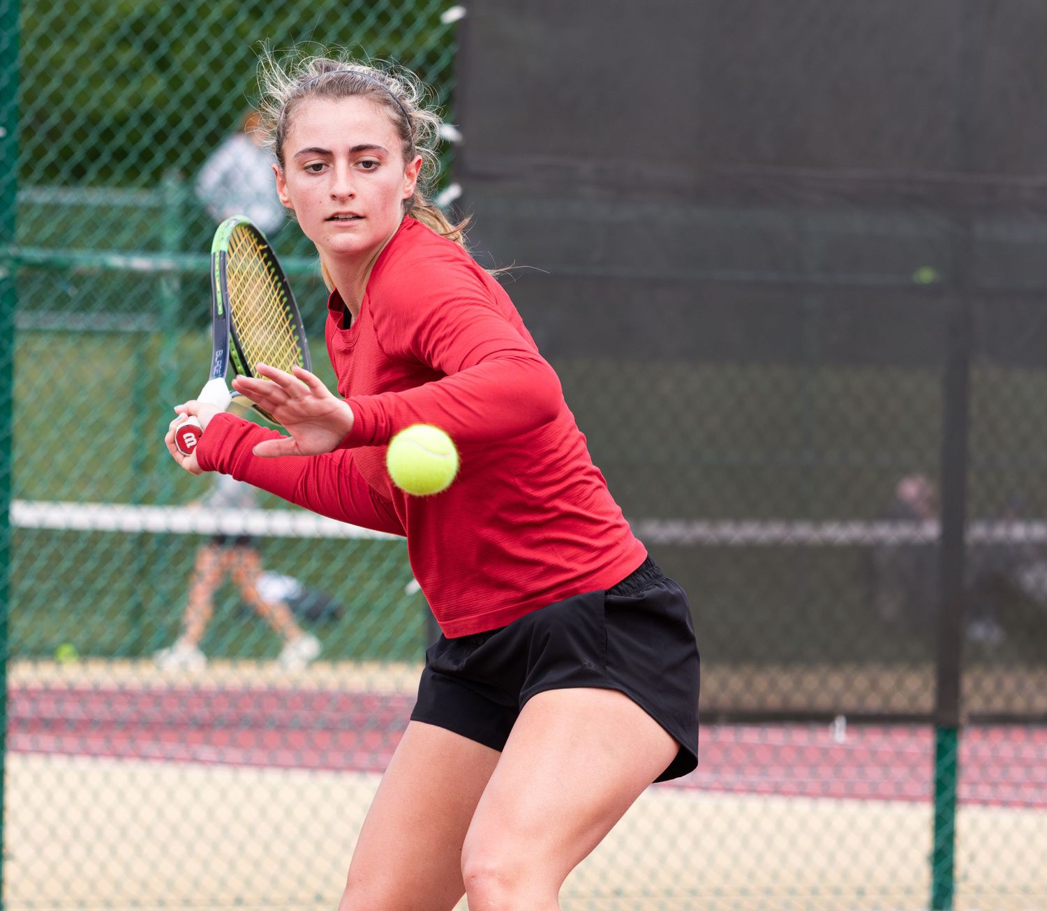 Tennis player in red shirt hitting forehand shot with yellow ball in motion.