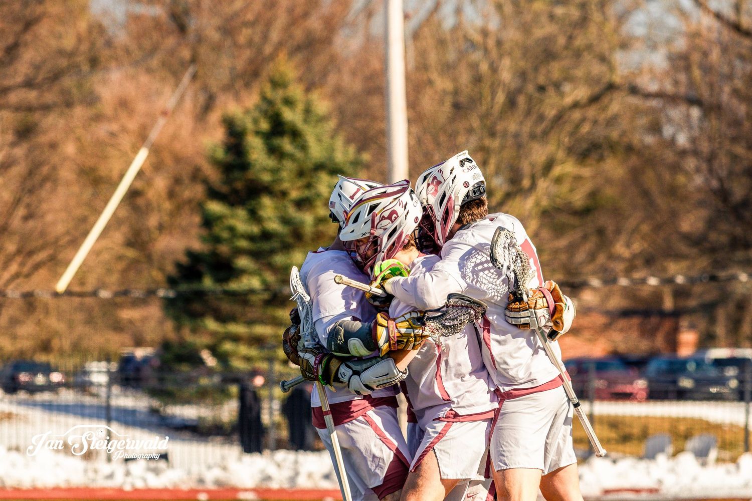 Lacrosse players huddle together on a snowy field during a winter game with trees visible in the background.