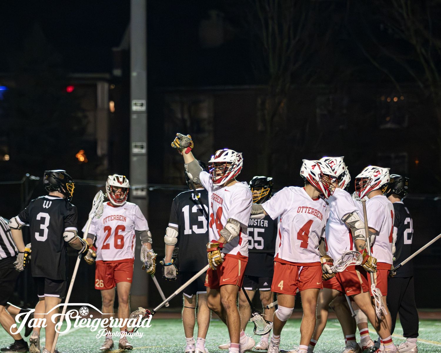 Lacrosse players in red and black uniforms celebrate on a floodlit field during a nighttime game.