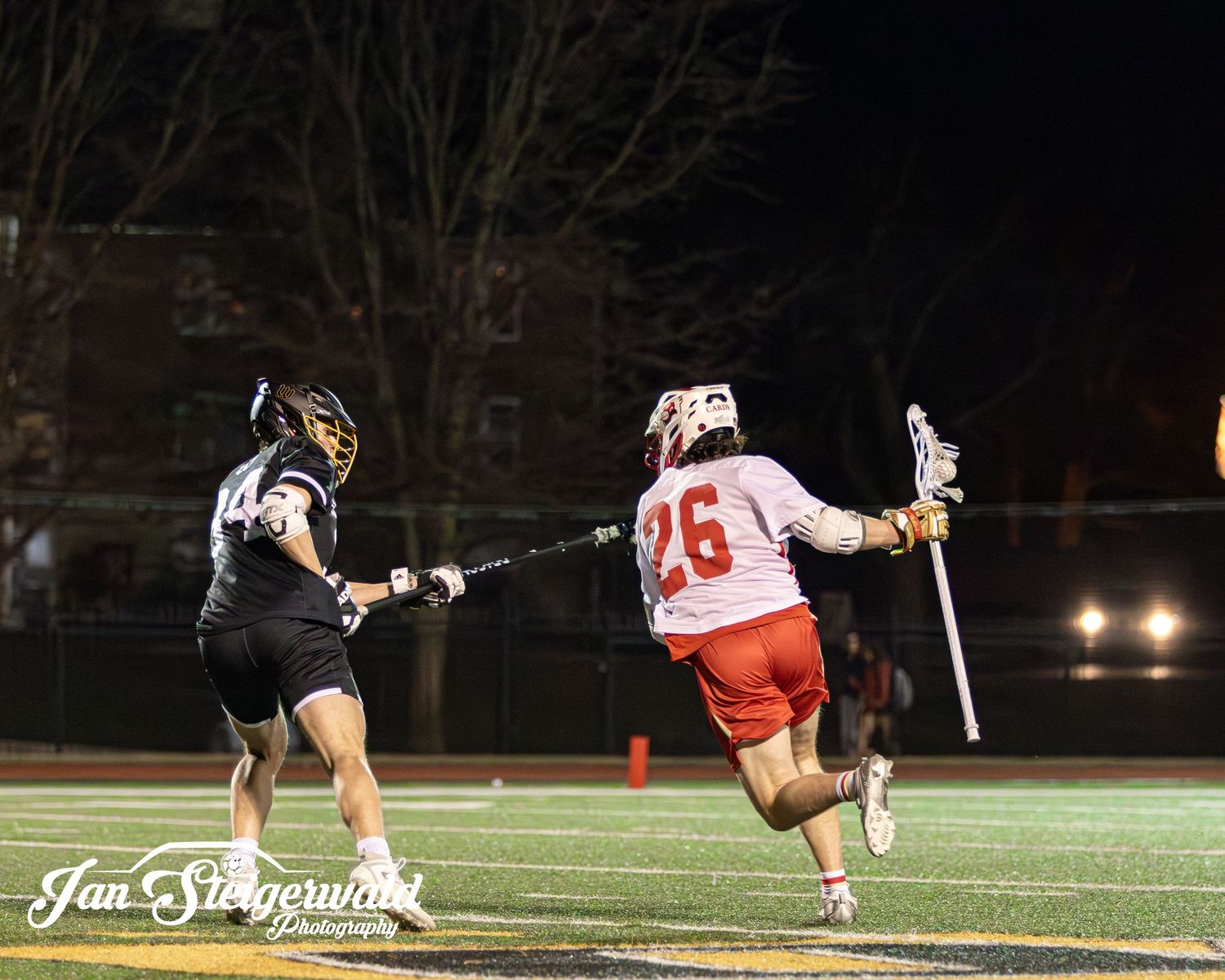 Lacrosse player in white jersey takes a diving shot during a nighttime game under stadium lights.