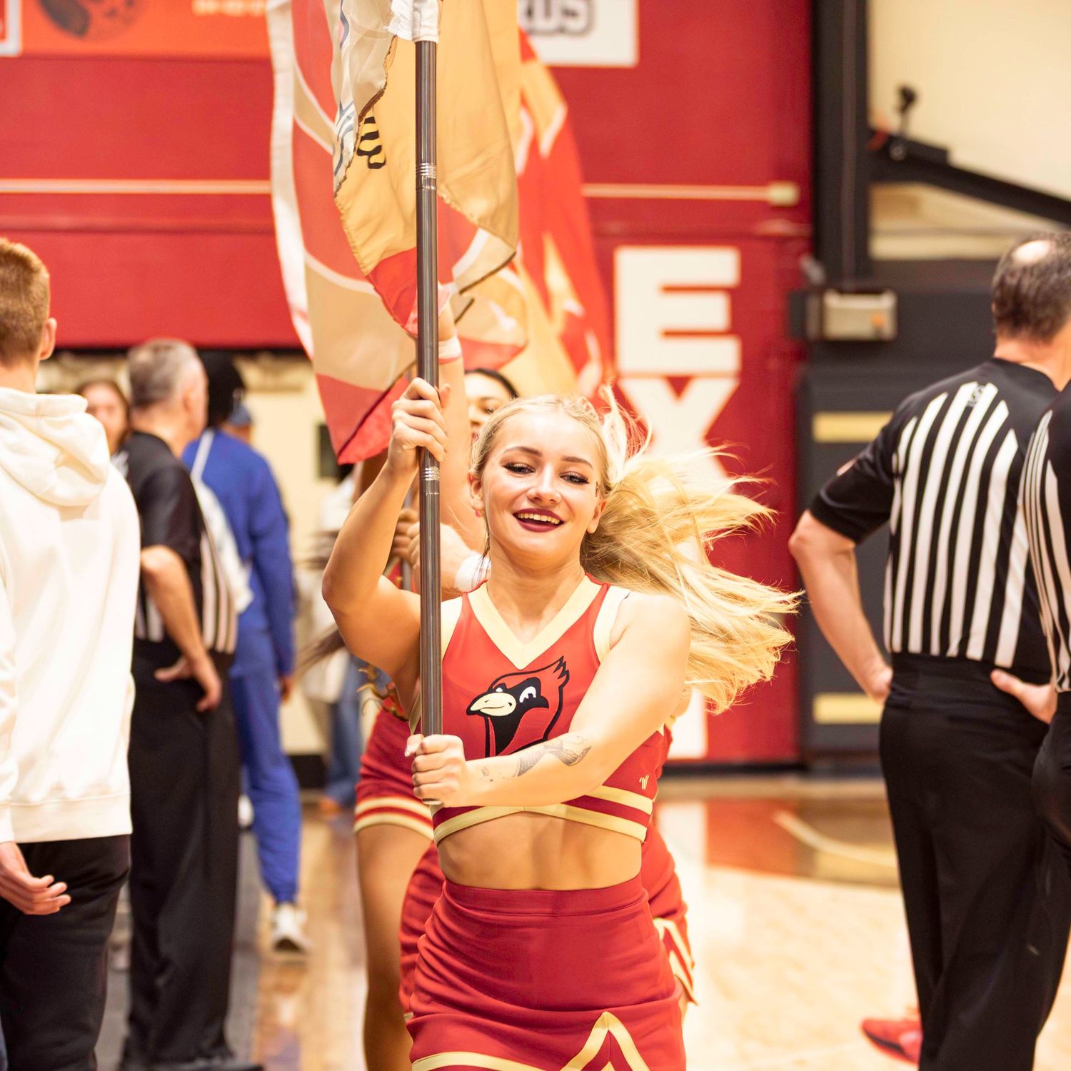 Cheerleader in red uniform performing with flag pole at indoor sporting event.