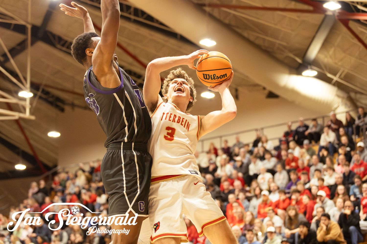 A basketball player jumps to make a shot during an intense game while spectators watch from the crowded stands.