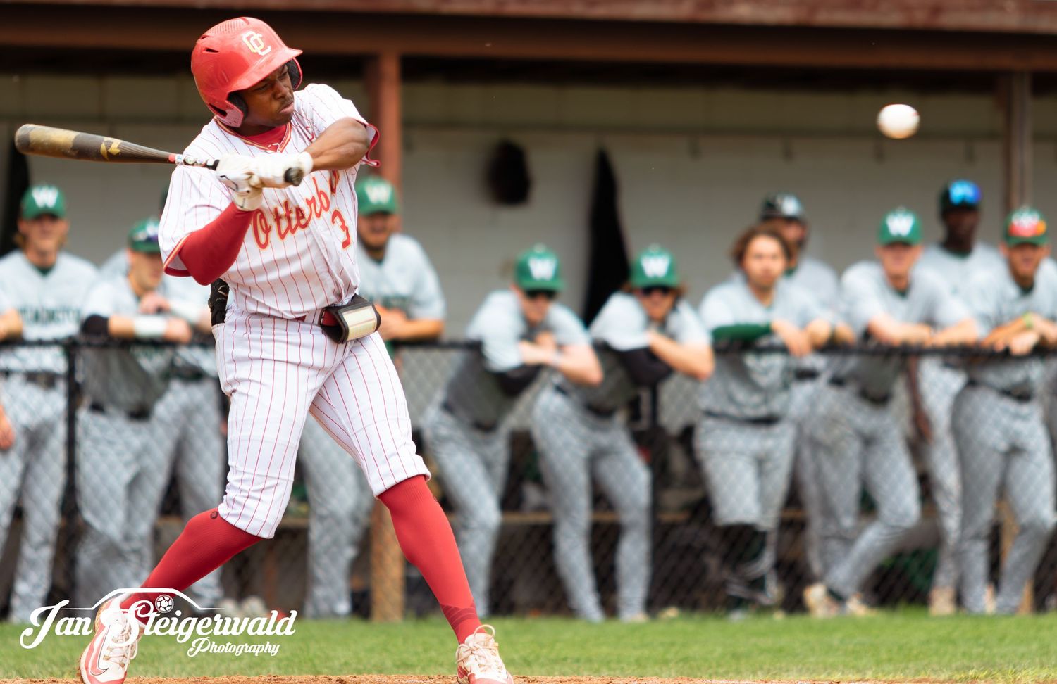 A baseball player in a white and red striped uniform swings at a pitch during a game as opponents watch from the dugout.