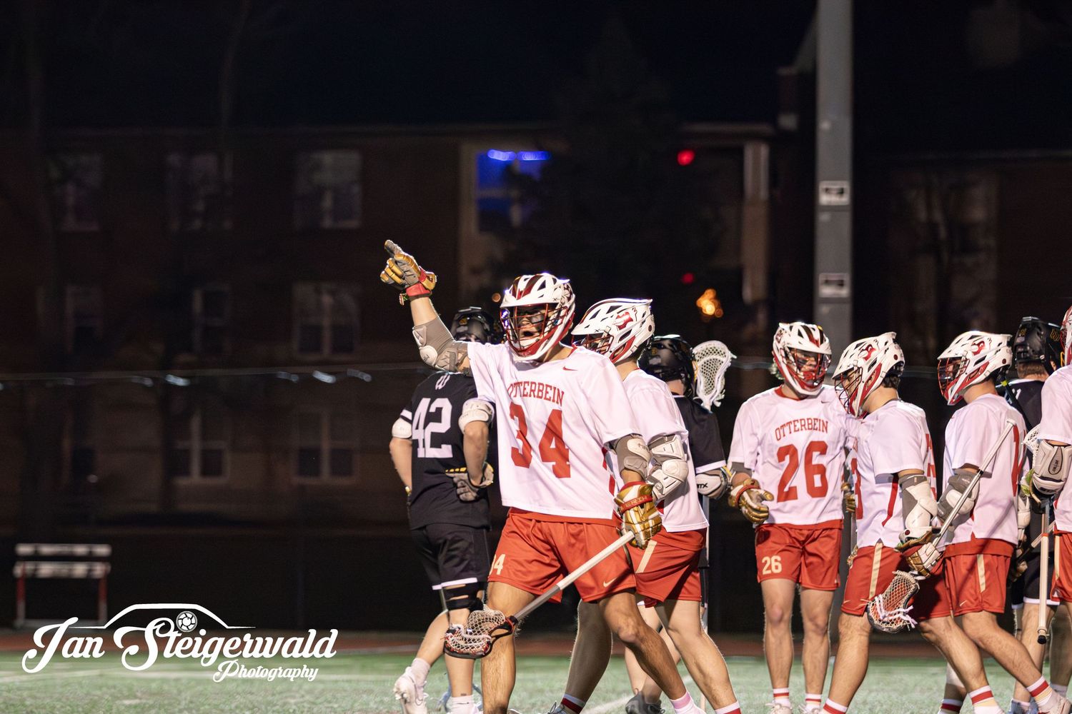 Lacrosse team players in white and orange uniforms celebrate together during a nighttime game under stadium lights.