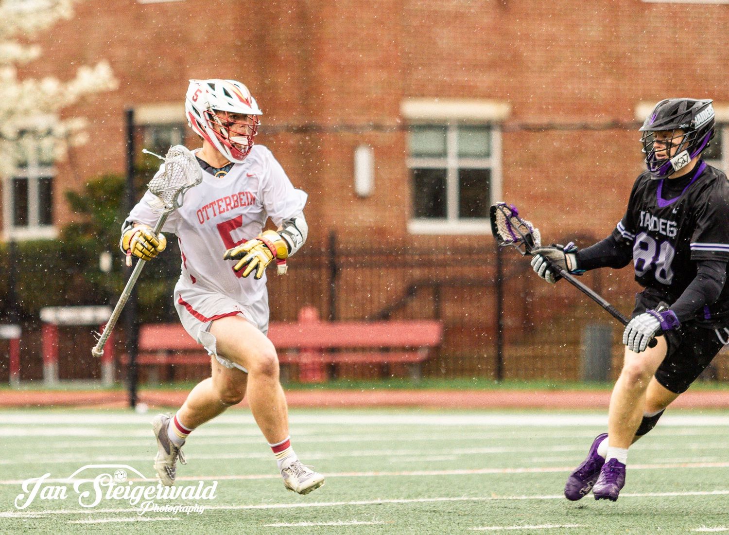 Two lacrosse players compete on a grass field with a red building visible in the background.