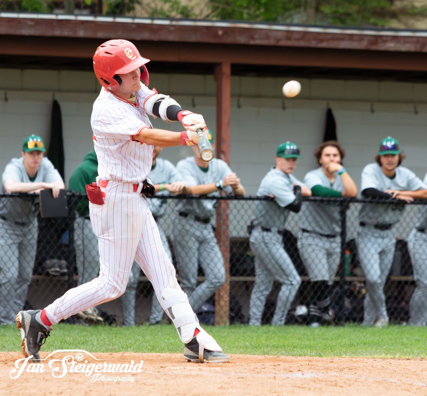 Baseball player in white pinstriped uniform swings bat to hit ball while opposing team watches from dugout.