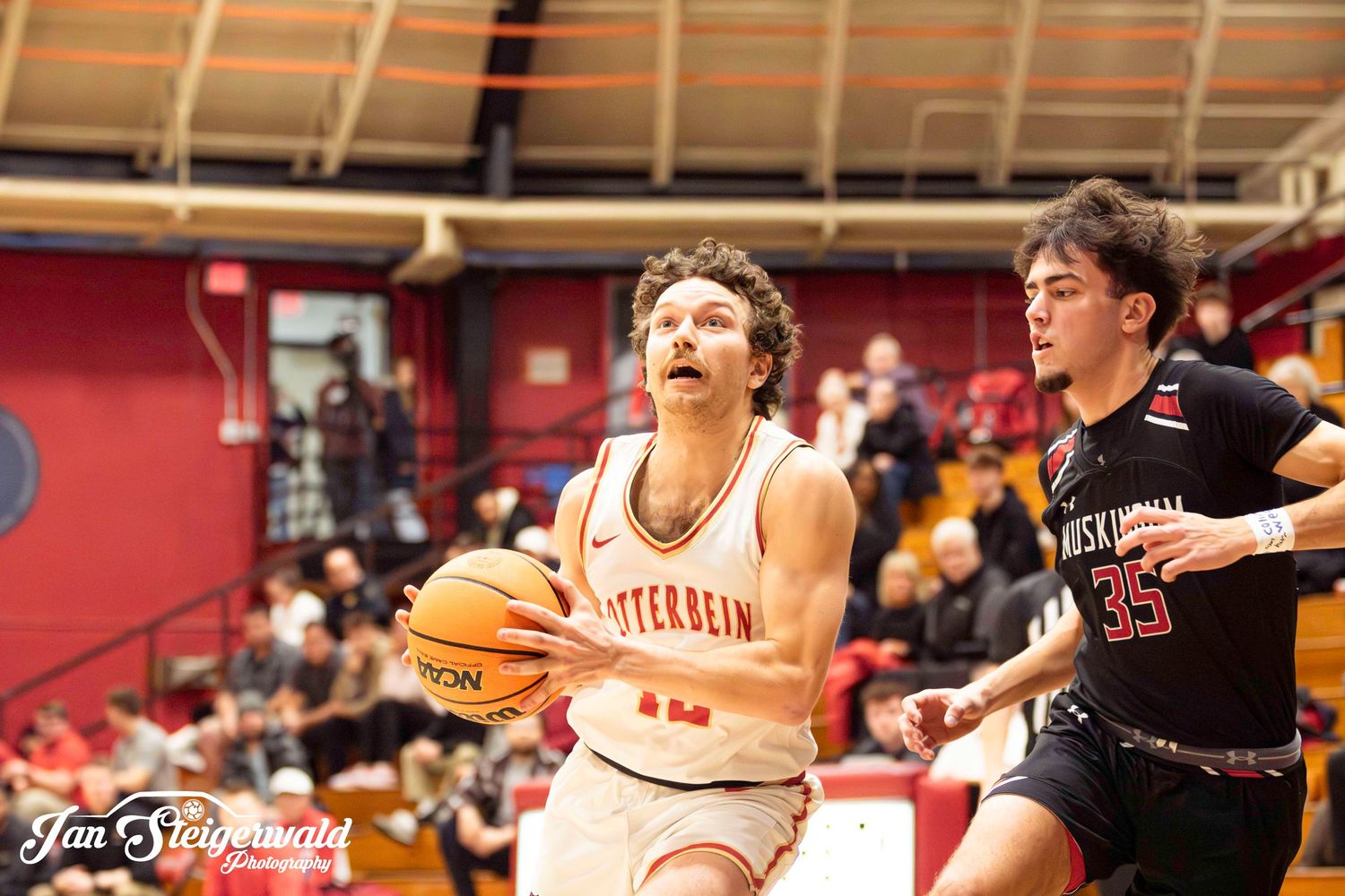 Basketball player drives towards basket while defender in black jersey pursues during indoor game.