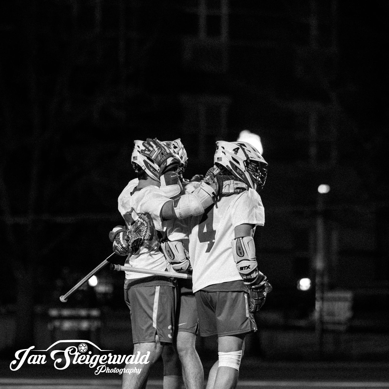 Two lacrosse players celebrate together during a nighttime game captured in black and white photography.