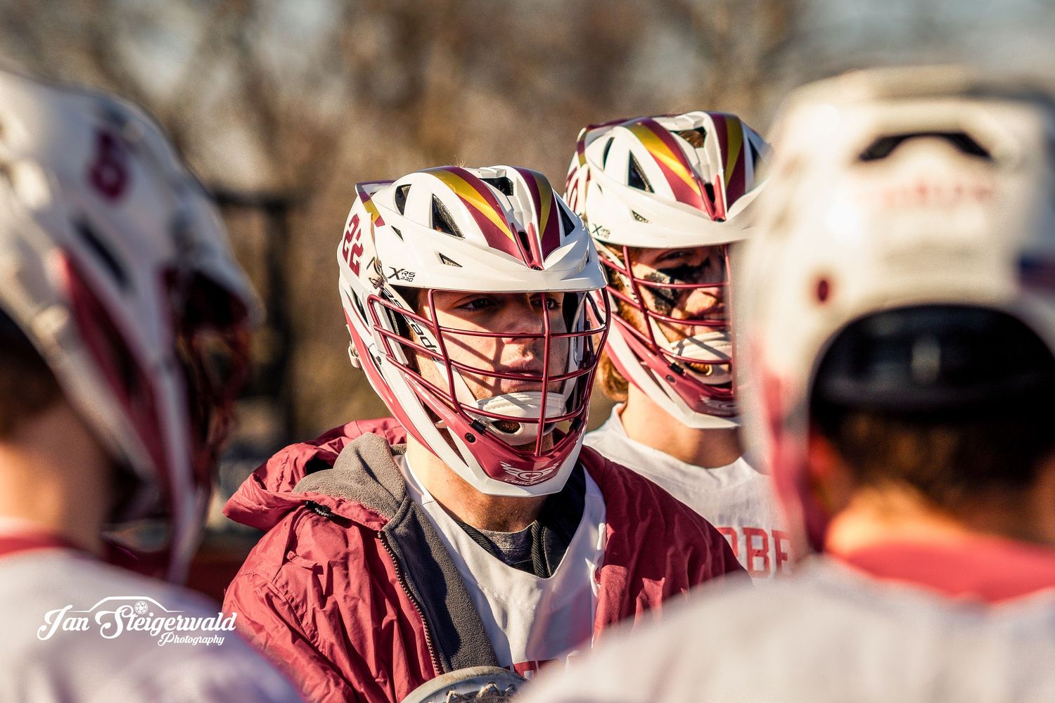 High school lacrosse players huddle together wearing red uniforms and white protective helmets during a game break.