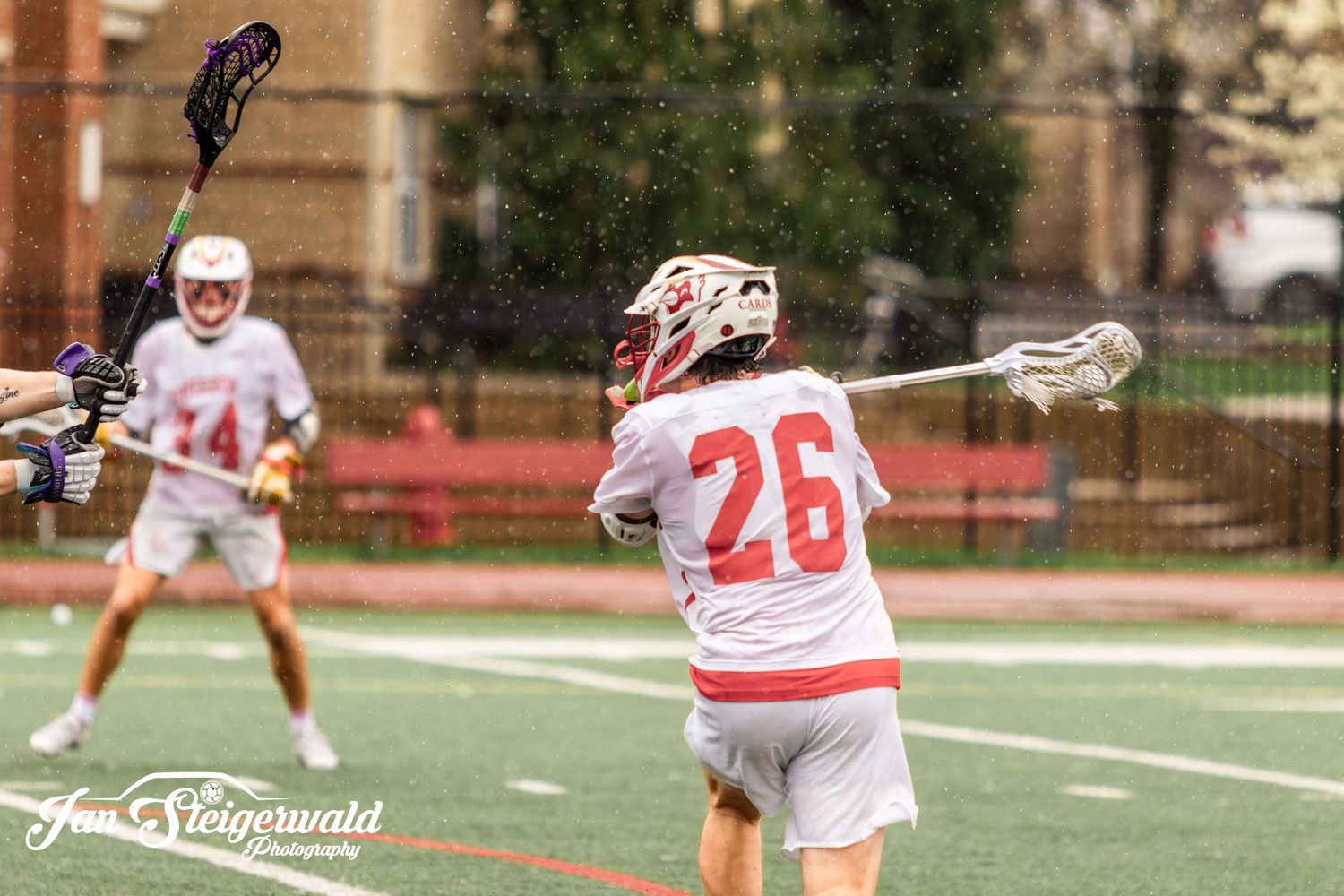Lacrosse player in white uniform number 26 cradles the ball while positioned on outdoor turf field.