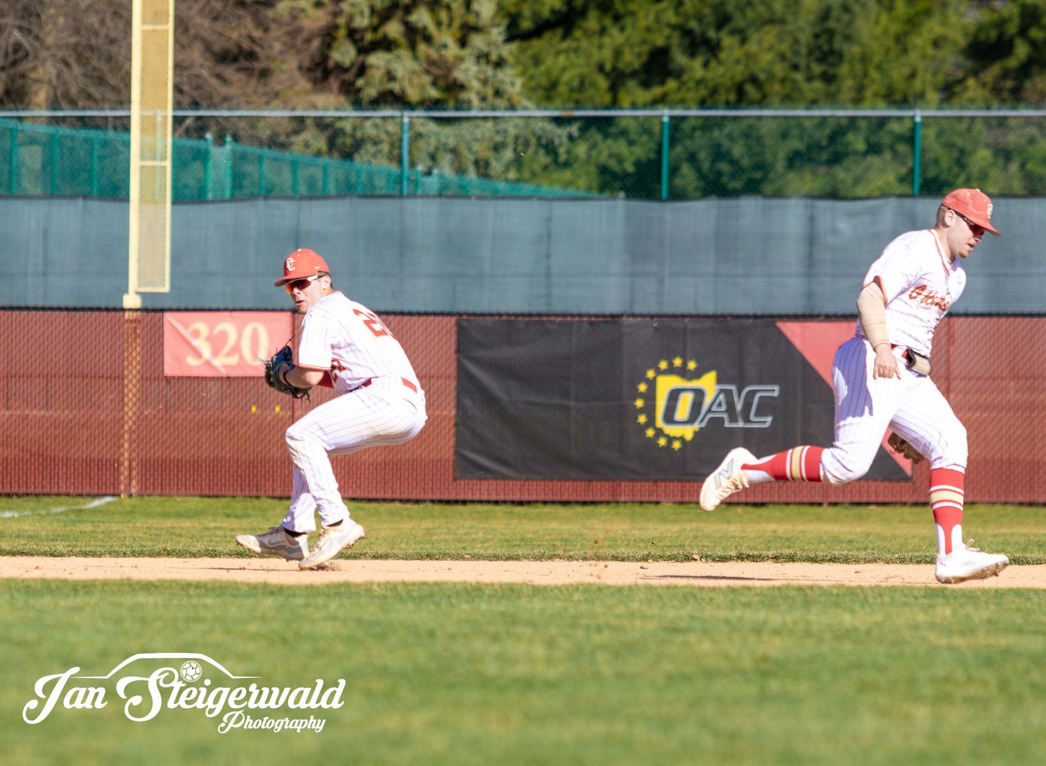 Baseball players in red uniforms lined up along a field fence during a game with green trees in the background.