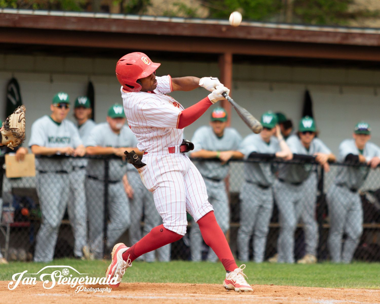 Baseball player in red and white pinstriped uniform swings bat during game with opposing team in background.