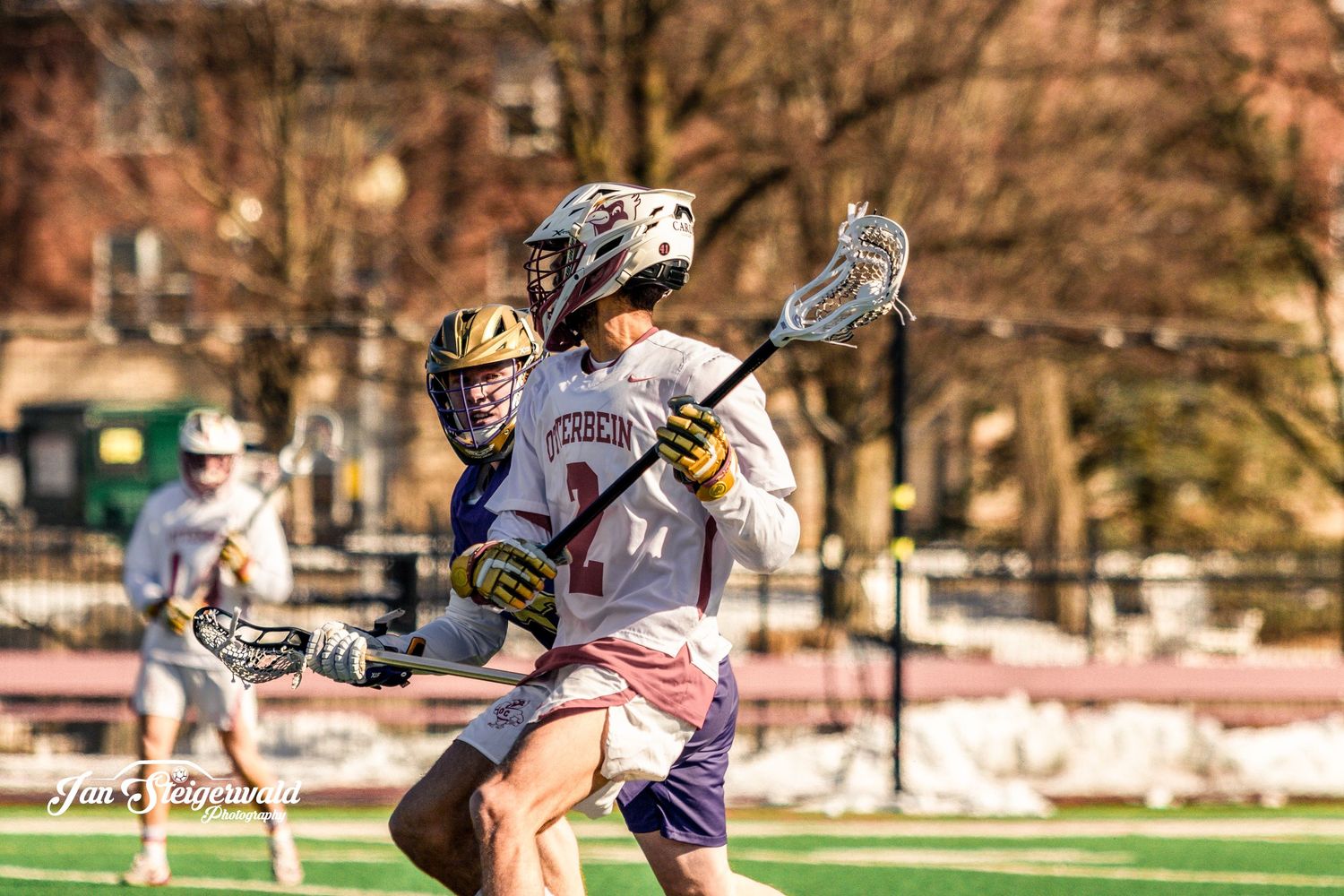 Lacrosse players in white and dark uniforms battle for possession during competitive match on outdoor field.