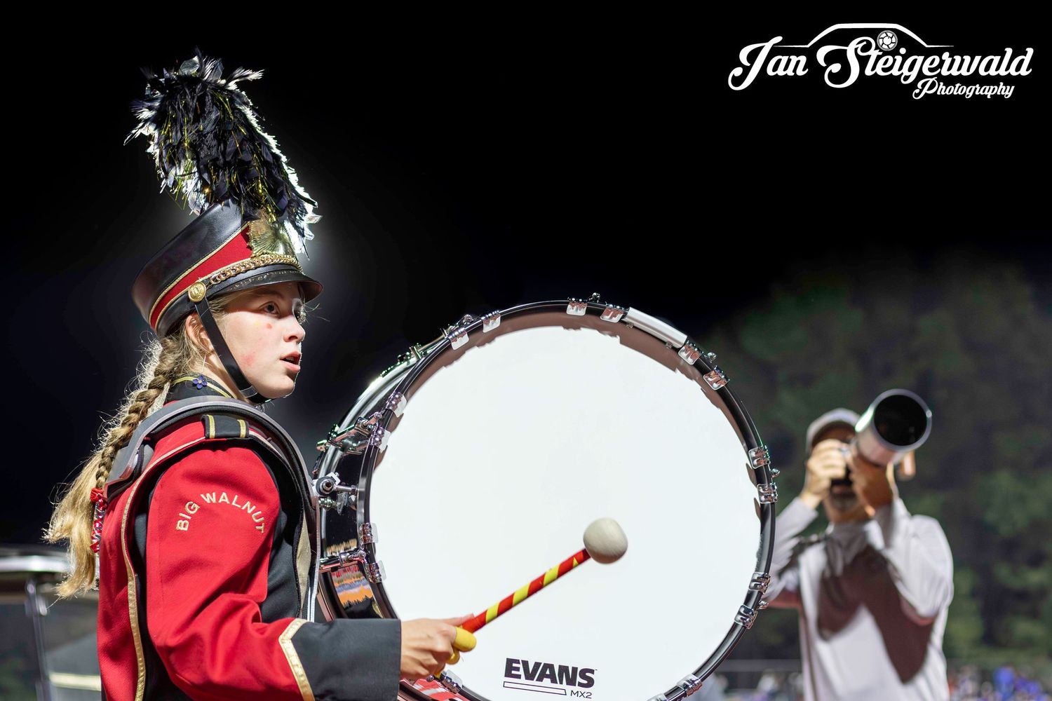 Marching band member in red uniform playing large bass drum during nighttime performance.