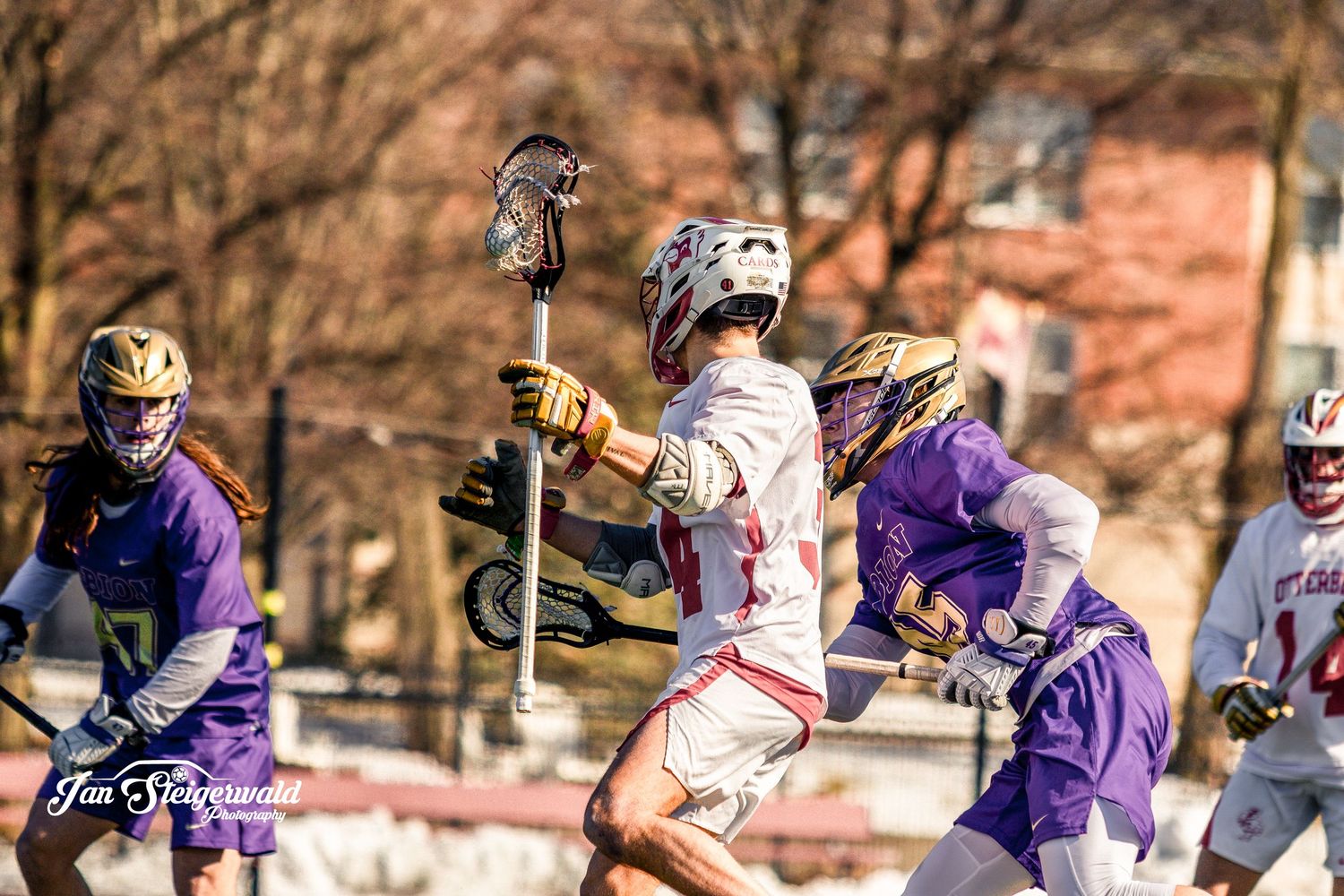 A college lacrosse game in progress with players from teams wearing purple and white uniforms competing on the field.