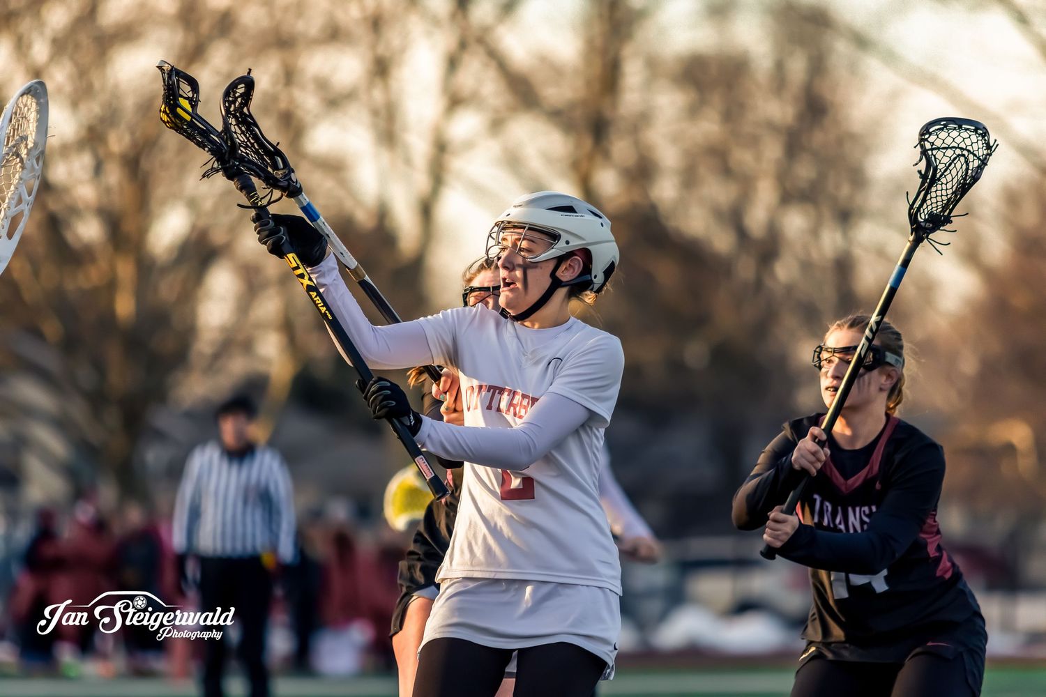 A lacrosse player in white uniform jumps with stick raised during a game against a defender at sunset.