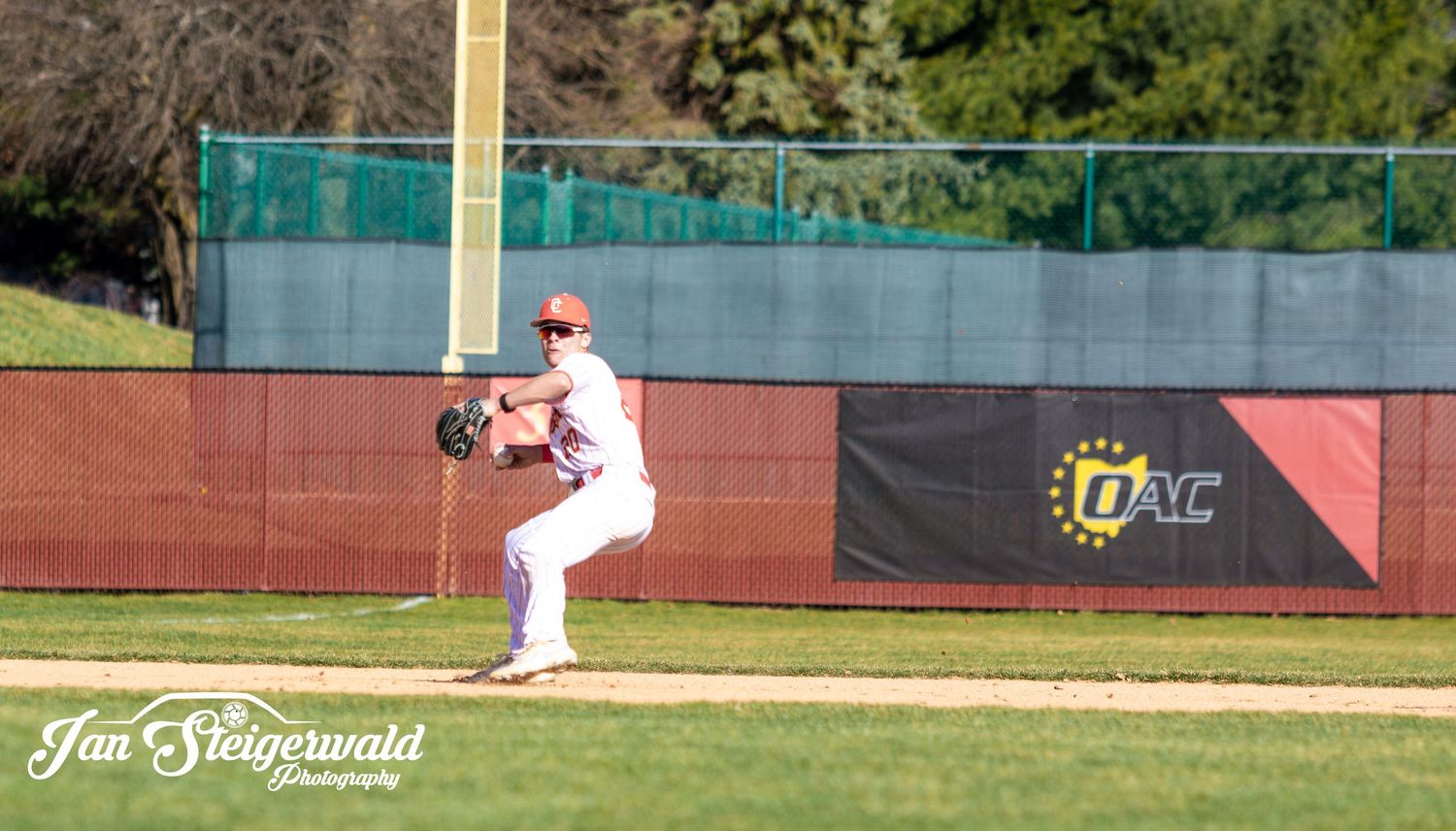 Baseball player in white uniform preparing to pitch on sunny day at baseball field with green fence in background.