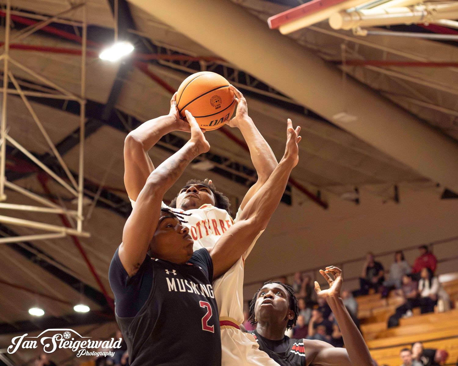 Basketball player in navy jersey goes up for a shot during an indoor game while being defended.