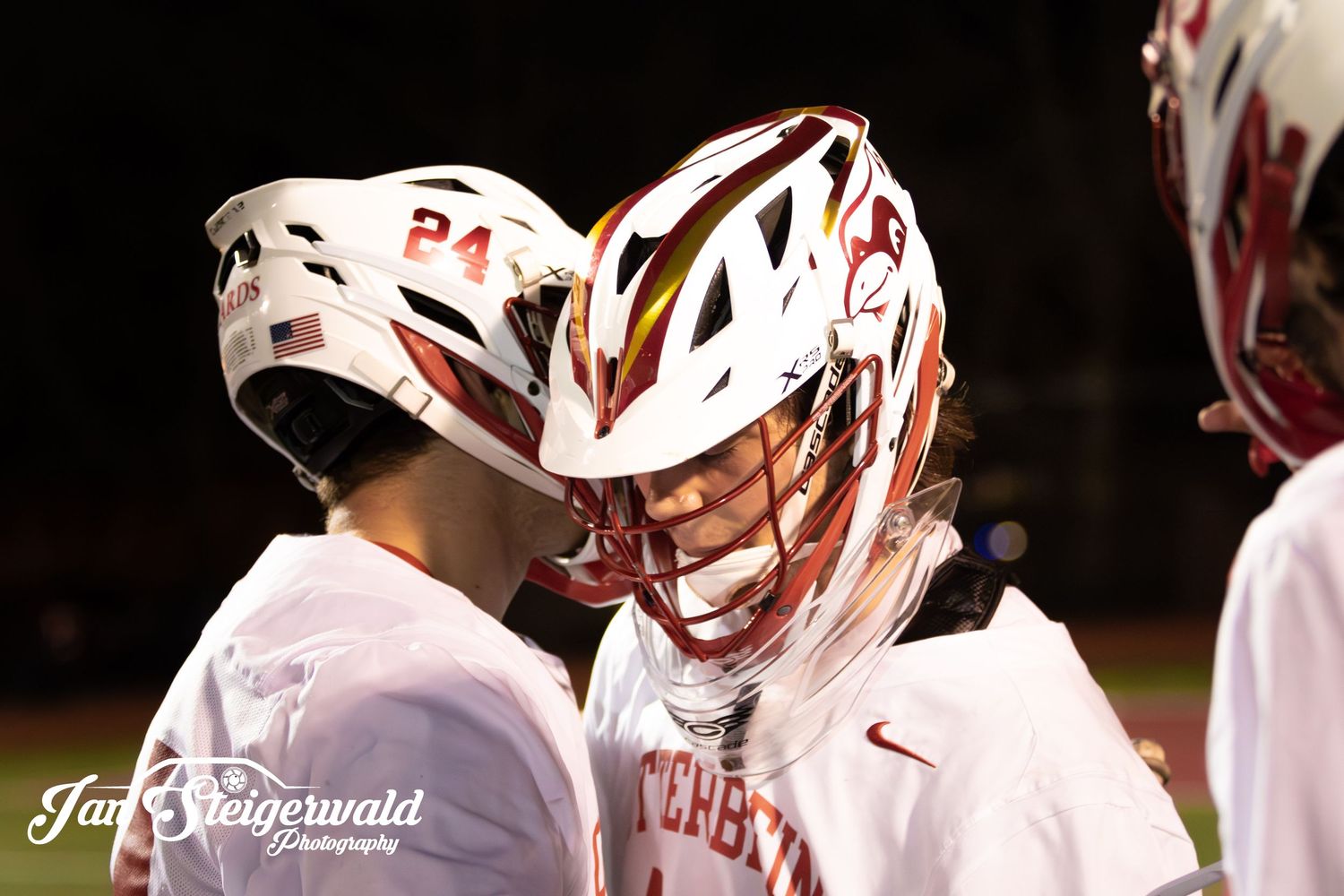 Two lacrosse players wearing white helmets and uniforms interact during a nighttime game.