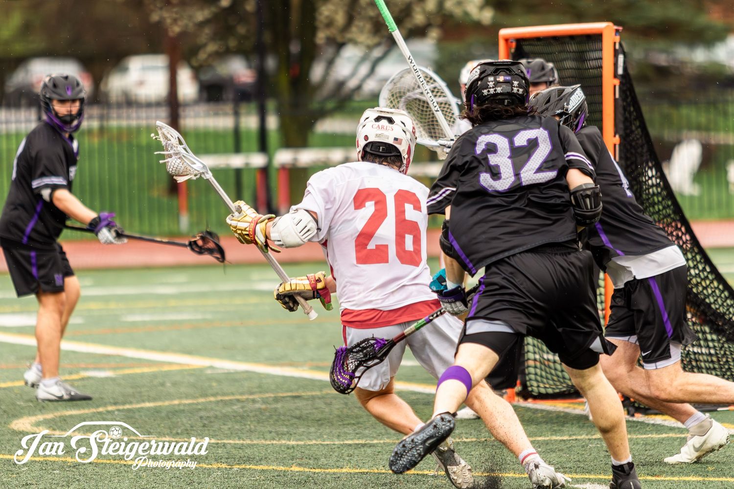 A fast-paced lacrosse game action shot shows players battling for position on a grass field during a match.